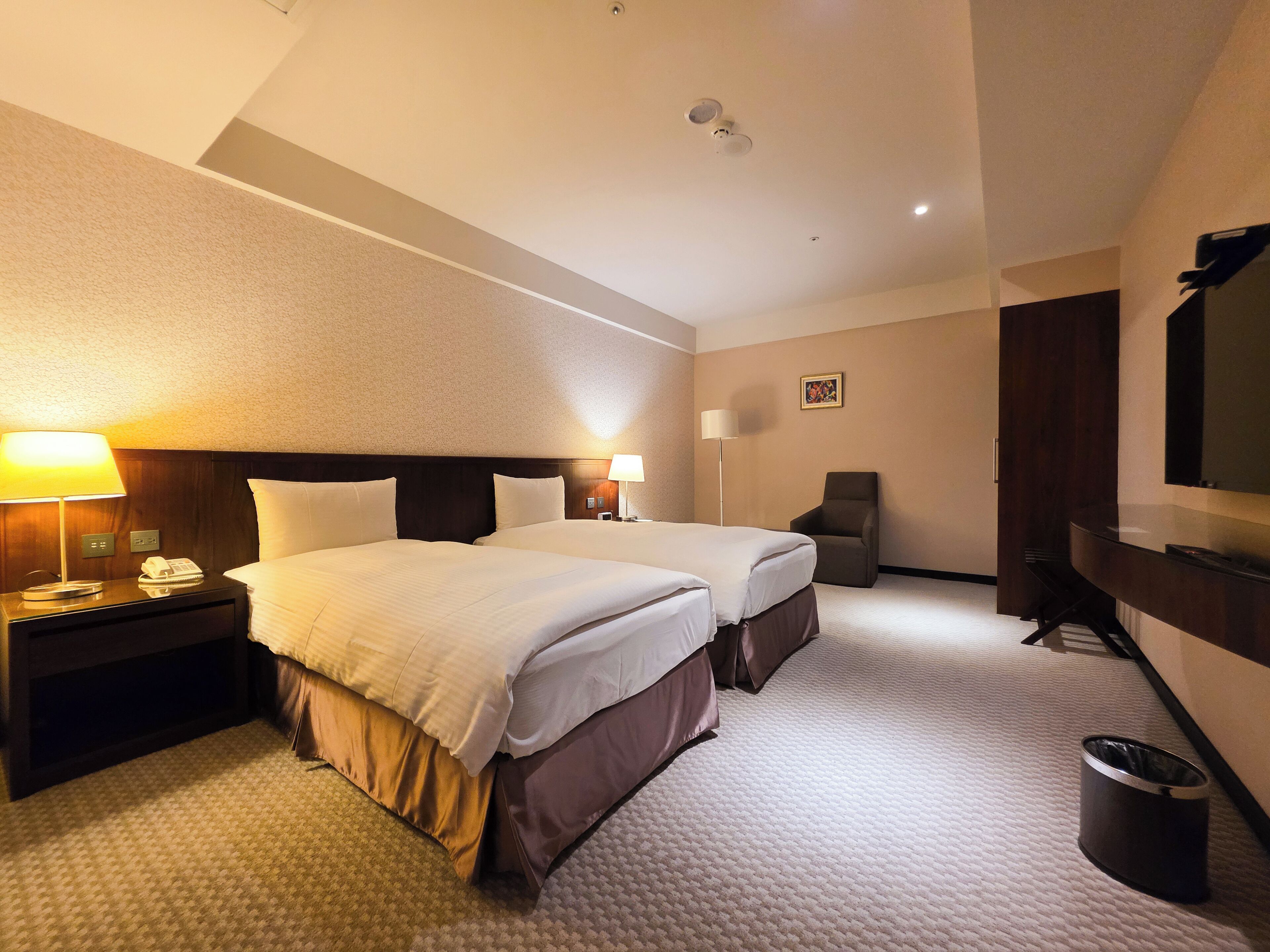 A hotel room with two beds, a desk with a lamp, a chair, a TV, and a trash can.