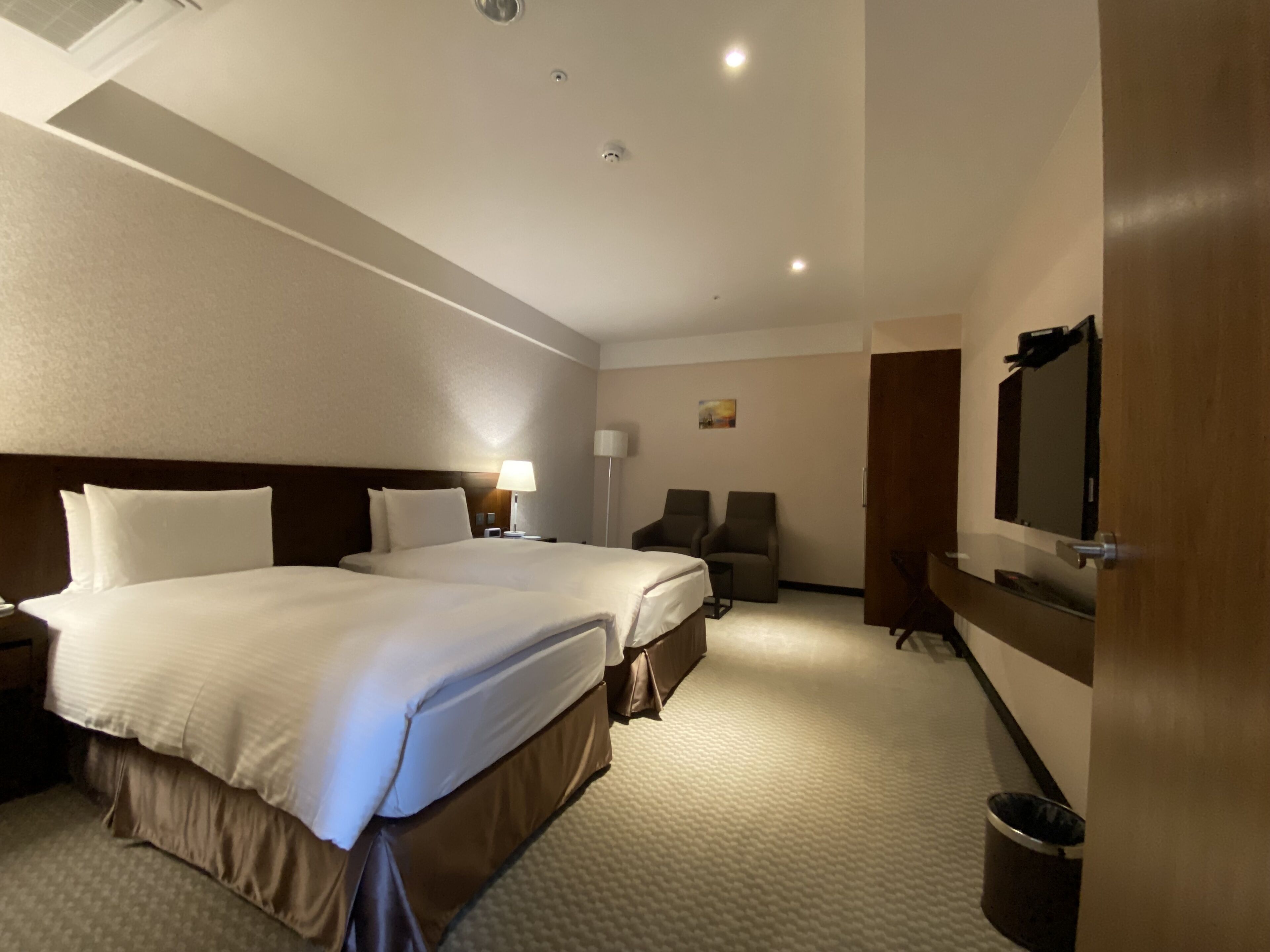 A hotel room with two beds, a desk, a chair, a TV, and a trash can.