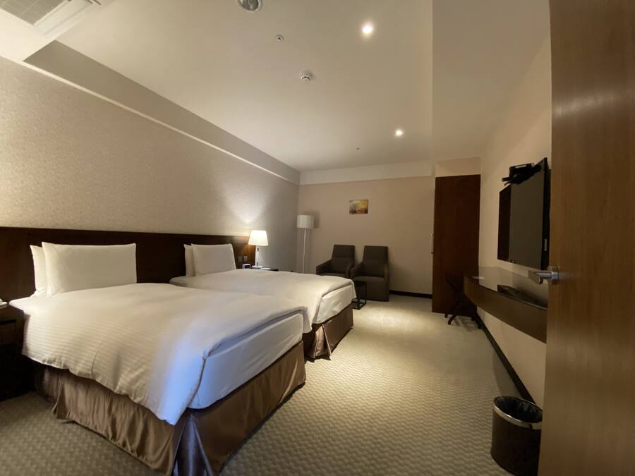 A hotel room with two beds, a desk, a chair, a TV, and a trash can.