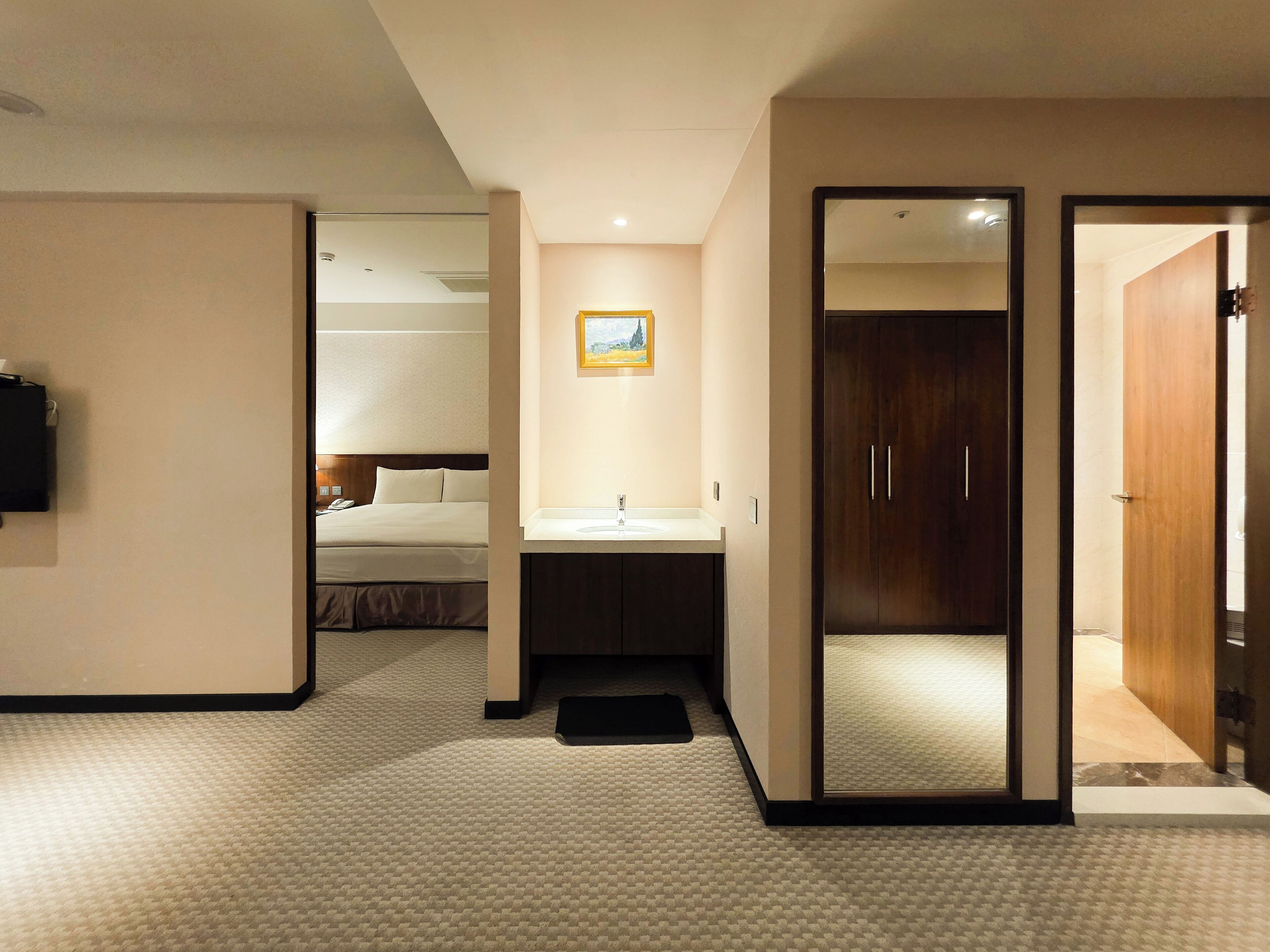 A hotel room with a bed, a bedside table, a mirror, a sink, and a door leading to another room.