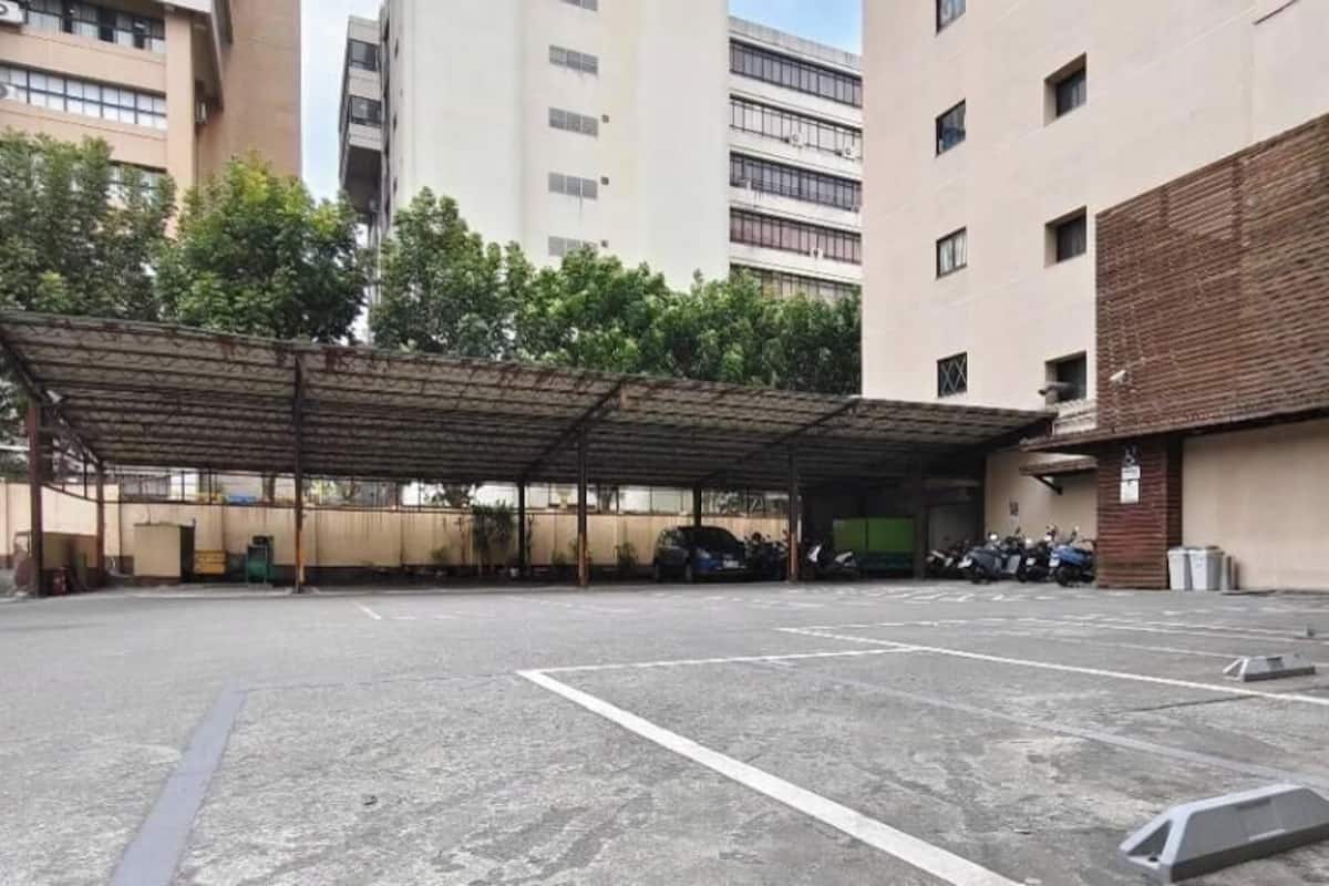 A parking area with a covered section, a building with a brick facade, and a multi-story structure in the background.