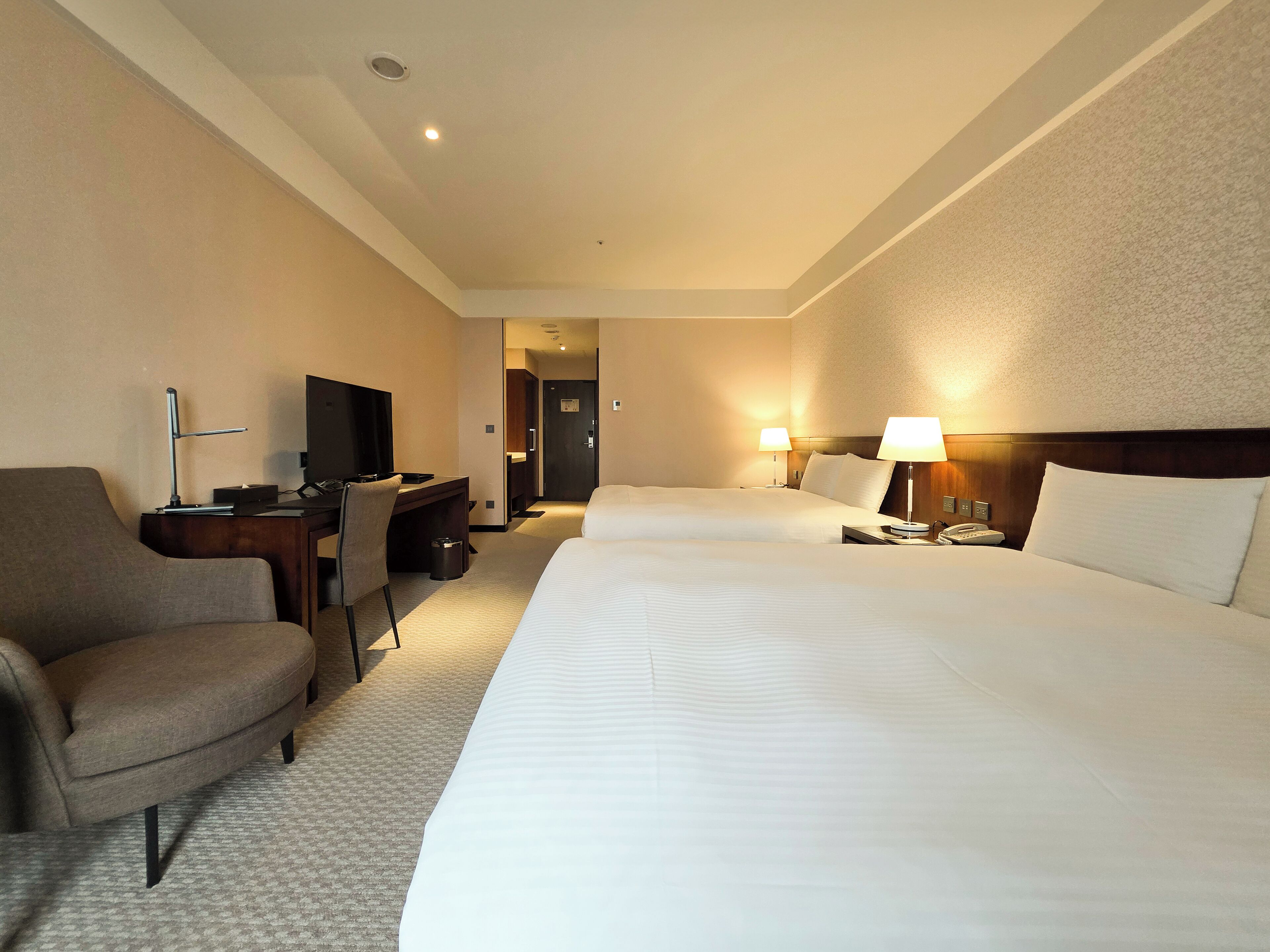 Quarto de hotel com uma cama grande, uma escrivaninha com cadeira, uma televisão e duas luminárias de cabeceira.