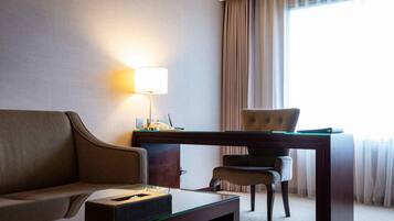 A hotel room with a sofa, a desk with a lamp, a chair, and a view of the outdoors through a window.
