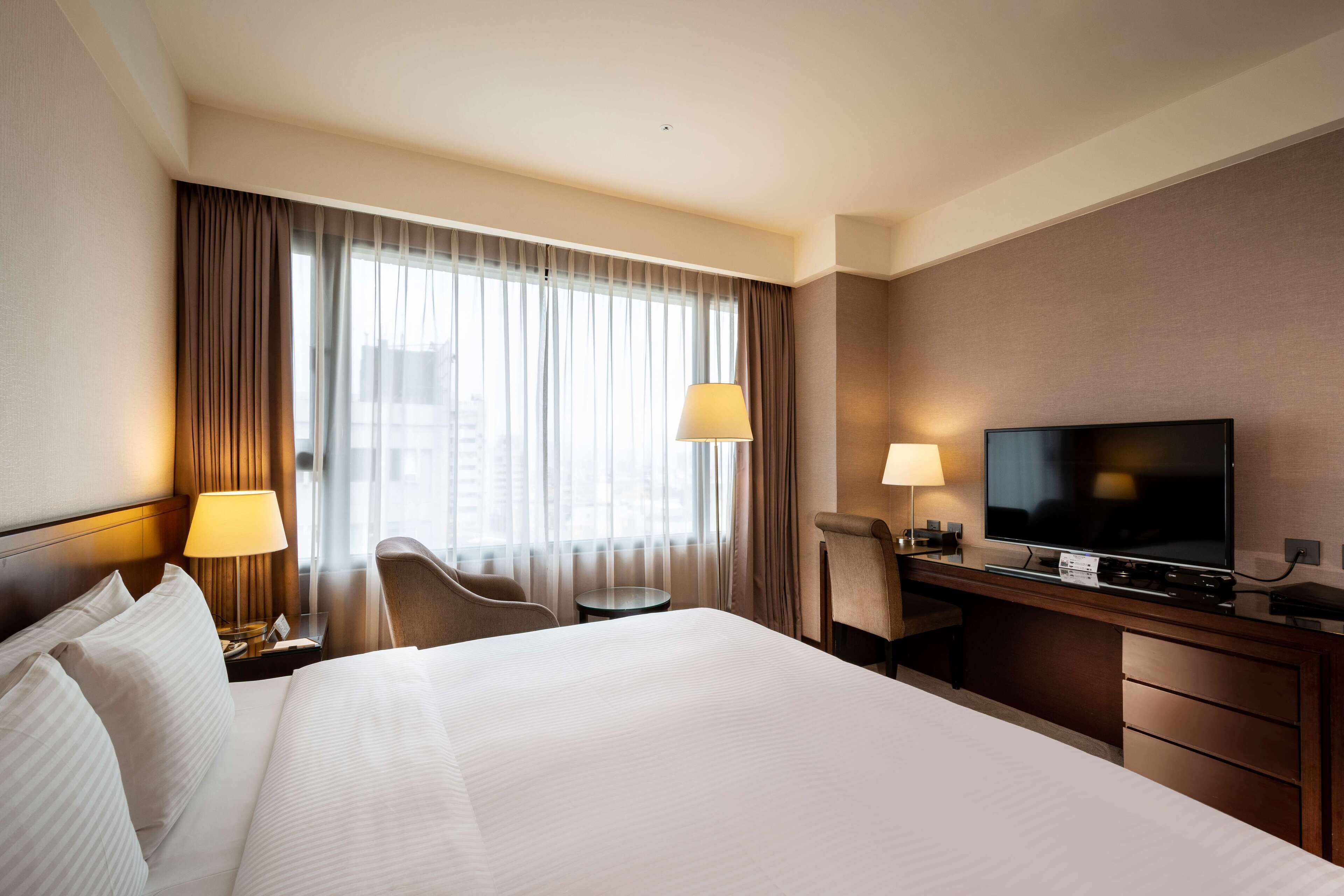 A hotel room with a large bed, a desk with a television, two lamps, and a chair.