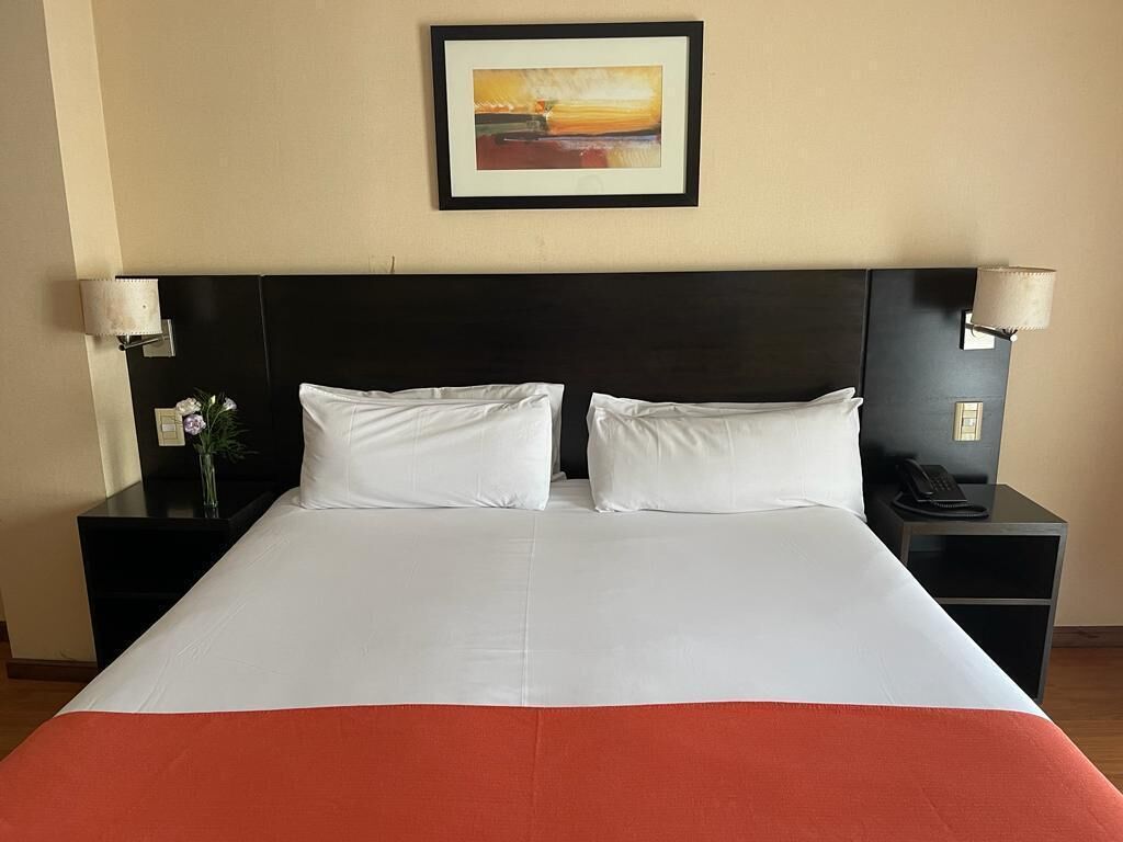 A hotel room with a large bed, two bedside tables, a nightstand, a framed picture on the wall, and a lamp on each bedside table.