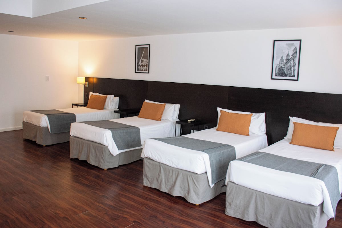 Une chambre d’hôtel avec quatre lits simples, chacun doté d’un couvre-lit gris et de draps blancs. On y trouve deux tables de chevet fixées au mur, avec des lampes, et le sol est recouvert de parquet.