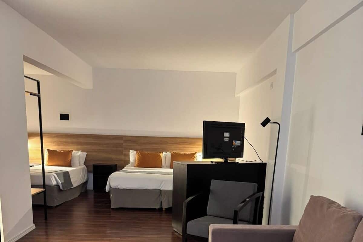 Une chambre d’hôtel moderne avec un plancher en bois, un canapé, un bureau avec un ordinateur et deux lits dotés d’une tête de lit en bois.