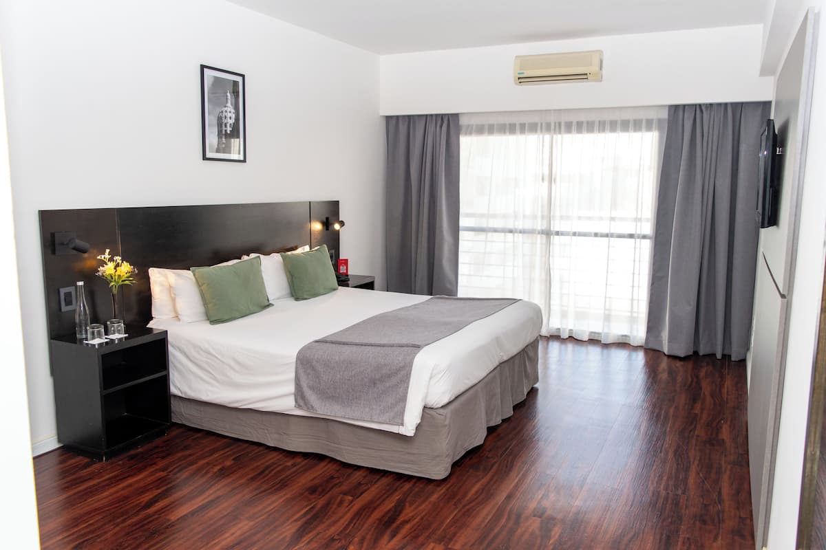 Une chambre d’hôtel bien rangée, avec un grand lit, une table de chevet avec une lampe, un tableau encadré au mur et une fenêtre avec des rideaux.