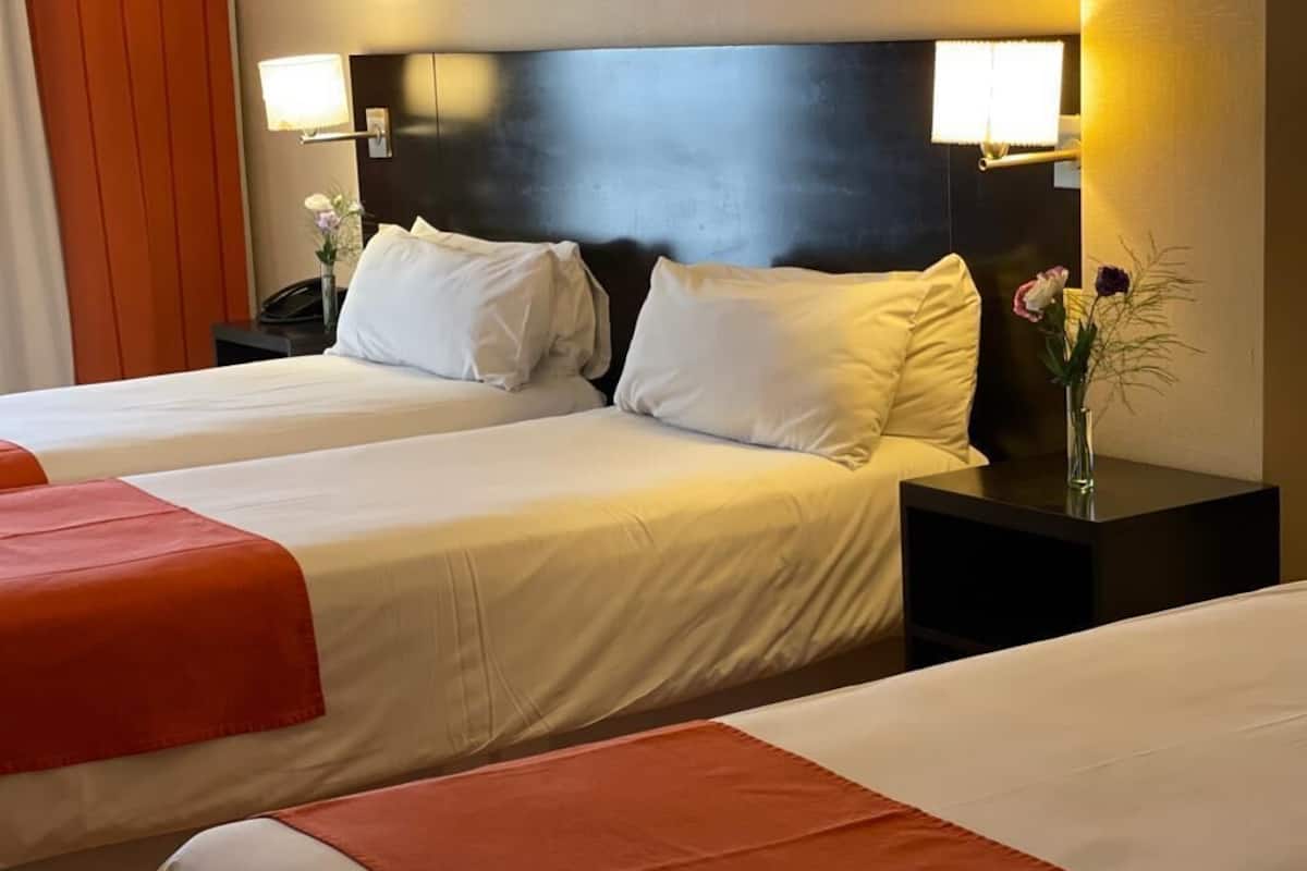Une chambre d’hôtel avec deux lits, chacun agrémenté de draps blancs et de coussins décoratifs orange. On y trouve des tables de chevet avec des lampes, et un vase rempli de fleurs sur l’une des tables.