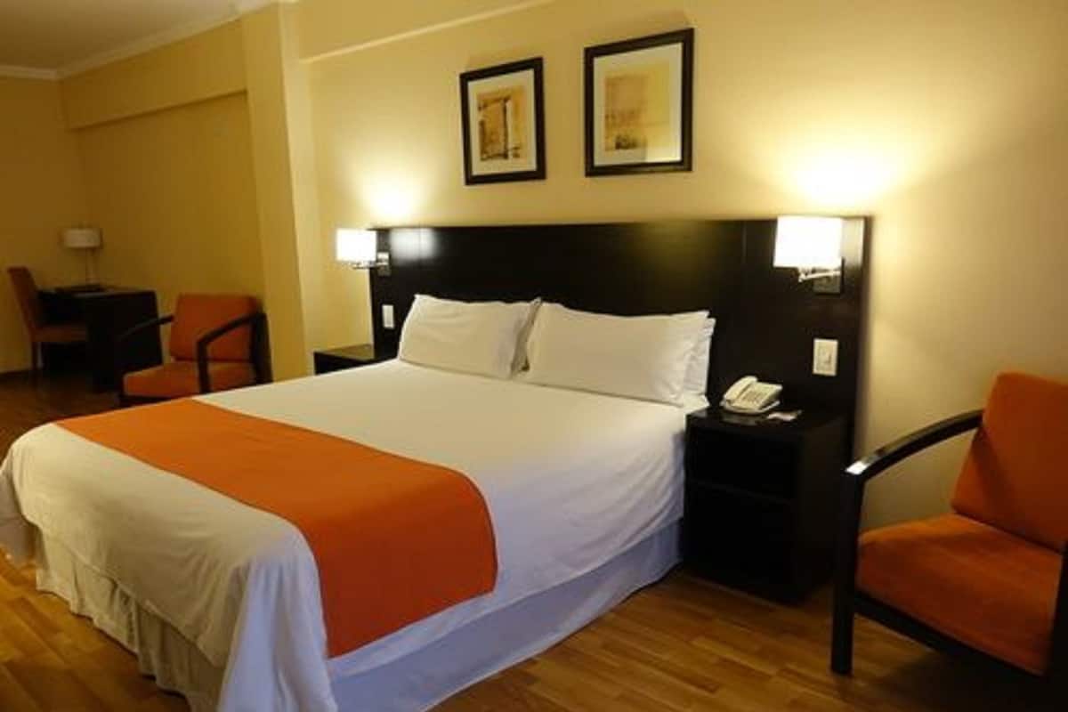 Une chambre d’hôtel avec un lit, deux chaises, une table de chevet avec un téléphone et deux tableaux encadrés au mur.