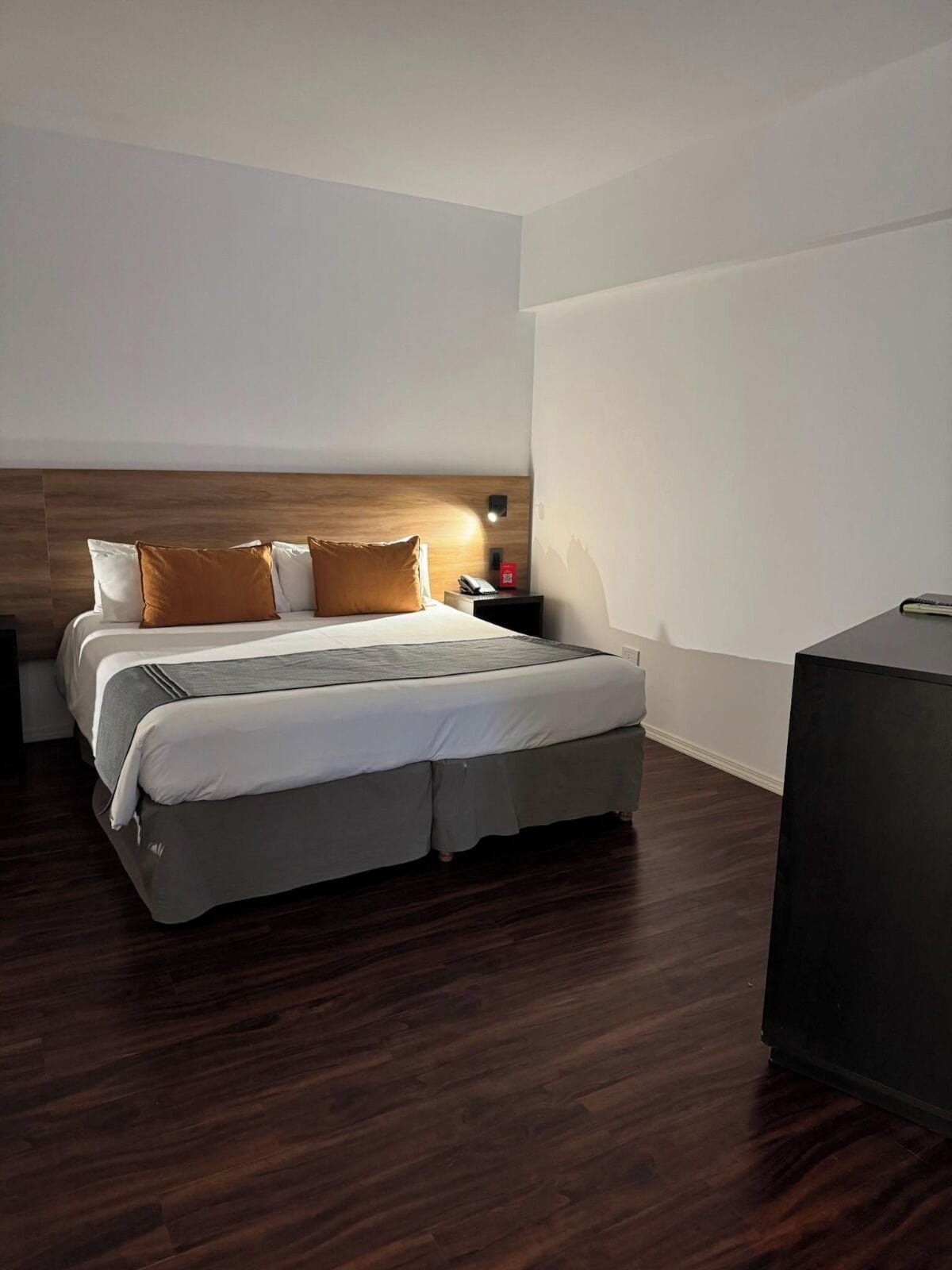 A modern hotel room with a wooden headboard, a neatly made bed with white and brown bedding, a bedside table with a lamp, and a dark wooden floor.
