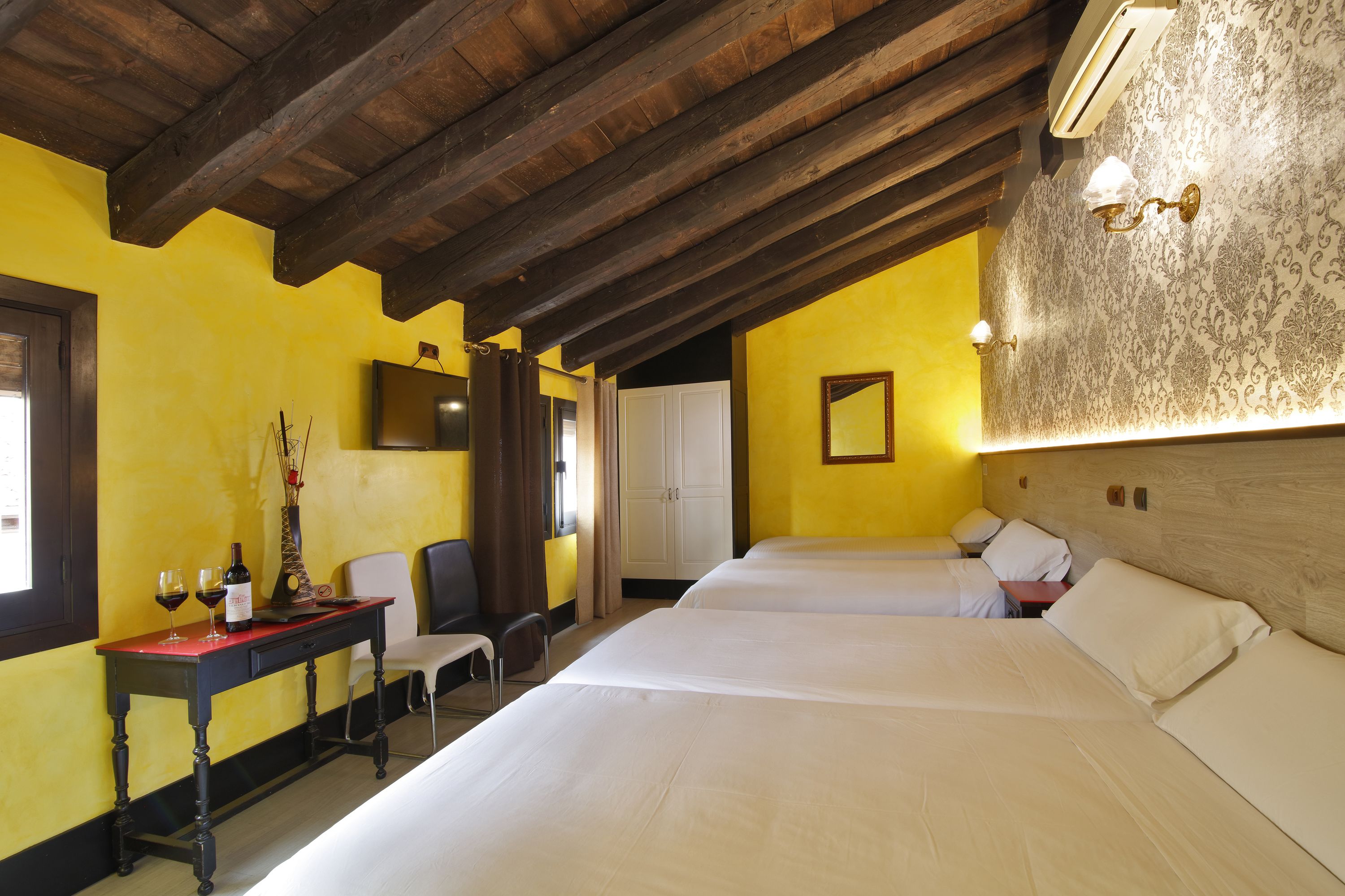 A room with two beds, a wooden ceiling, a desk with a chair, and a mirror on the wall.