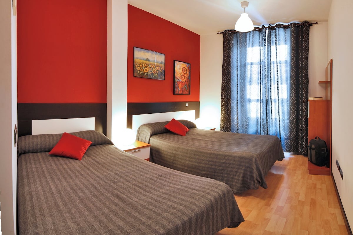 Een hotelkamer met twee bedden, een rode accentmuur, een schilderij en een raam met gordijnen met een patroon.