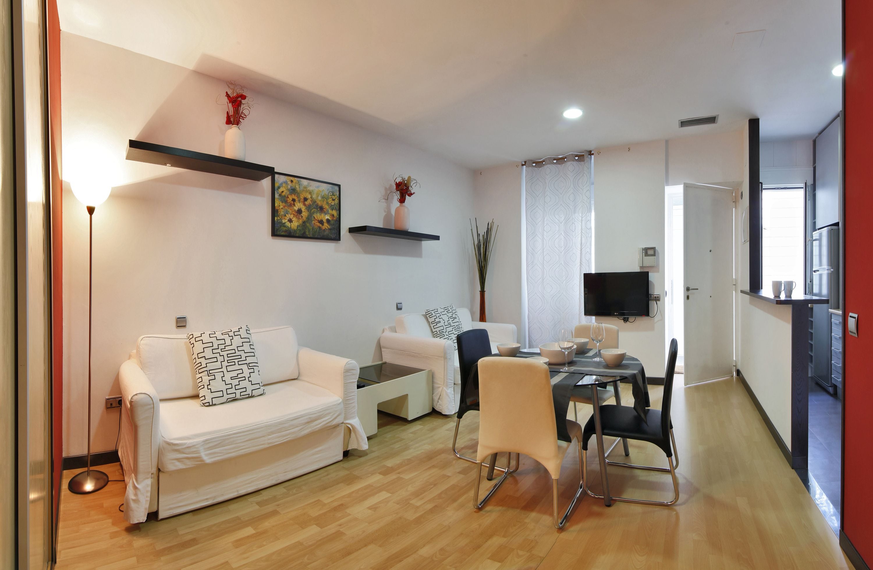 A modern living room with a sofa, coffee table, and dining area. There is a TV mounted on the wall and a kitchen area visible in the background.