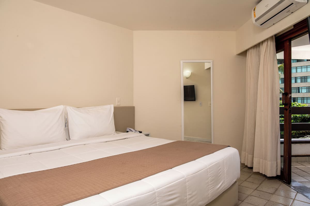 A hotel room with a bed, a bedside table, a mirror, and a view of the outside balcony.