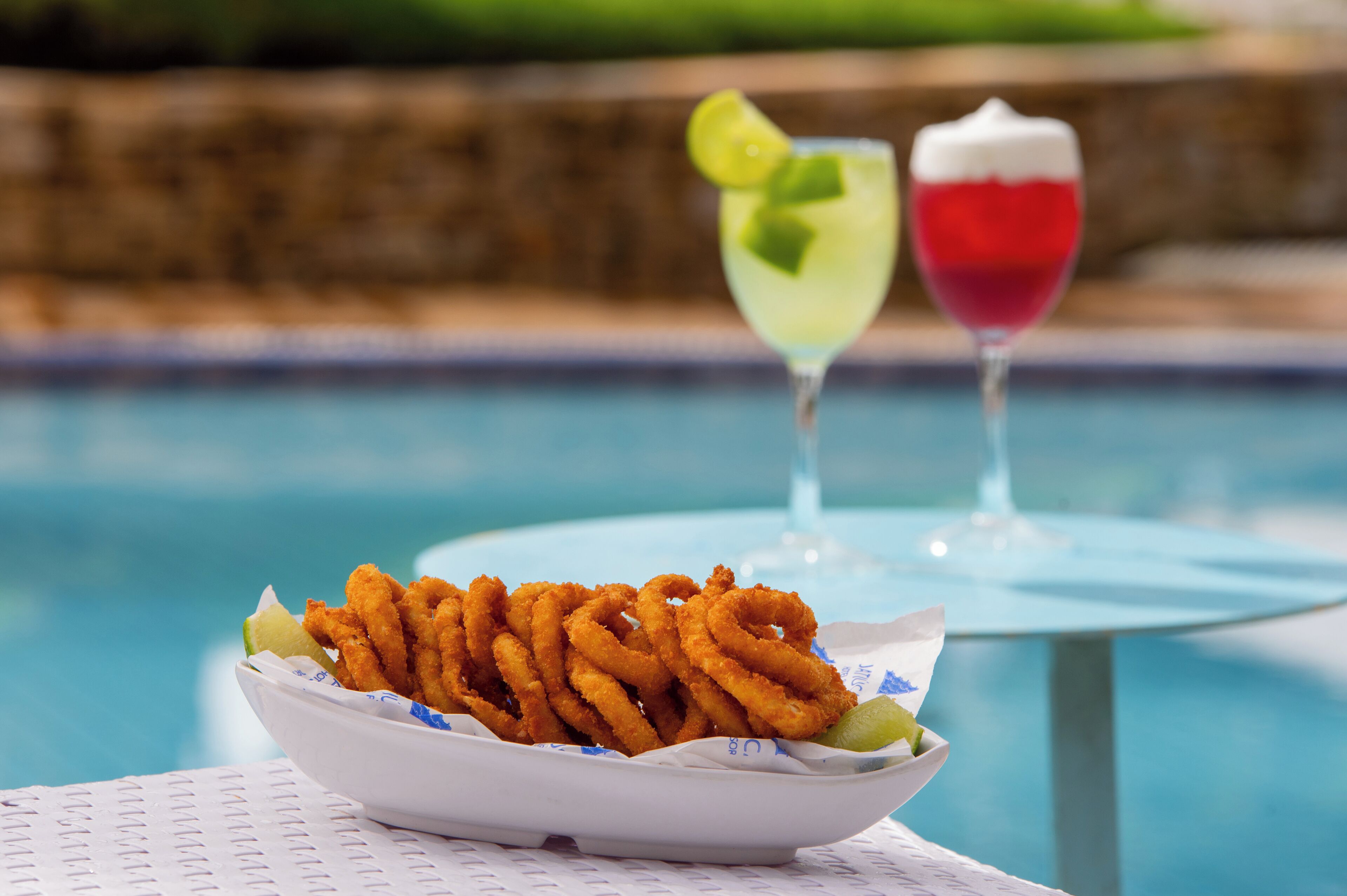 Un bol de rondelles d’oignon frites, accompagné d’un quartier de citron vert, et deux cocktails posés sur une table au bord d’une piscine.