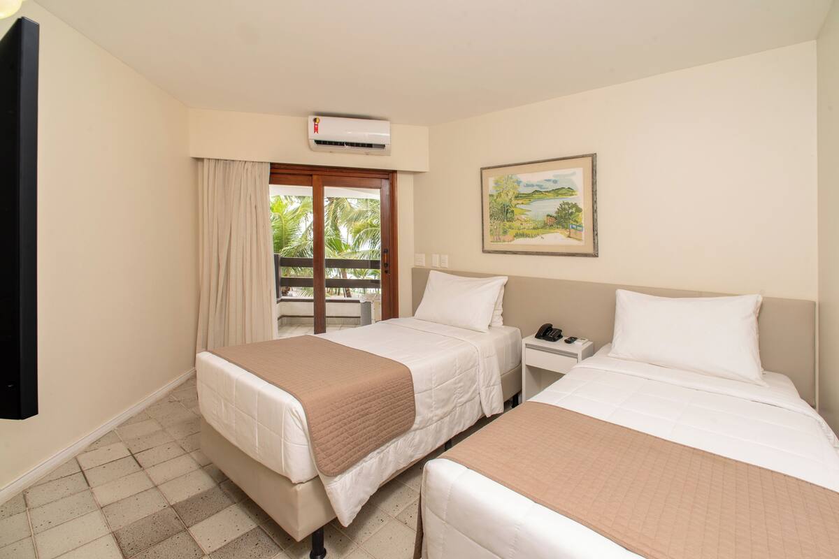 A hotel room with two beds, a flat-screen TV, an air conditioner, a painting, and a view of palm trees through a sliding door.