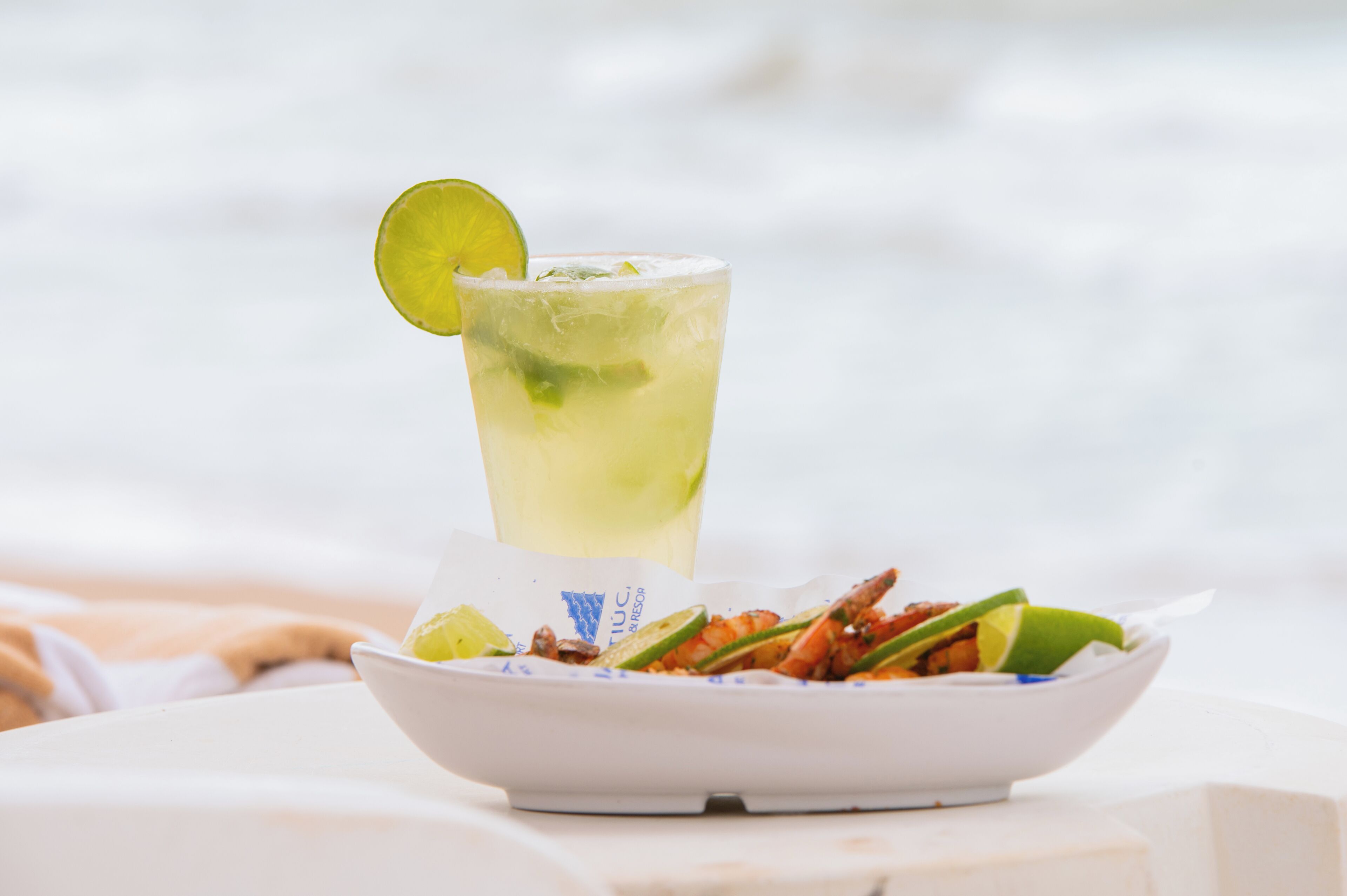 Un vaso de una bebida de color claro, adornado con una rodaja de lima, un plato de mariscos y una fuente blanca con un logotipo azul.