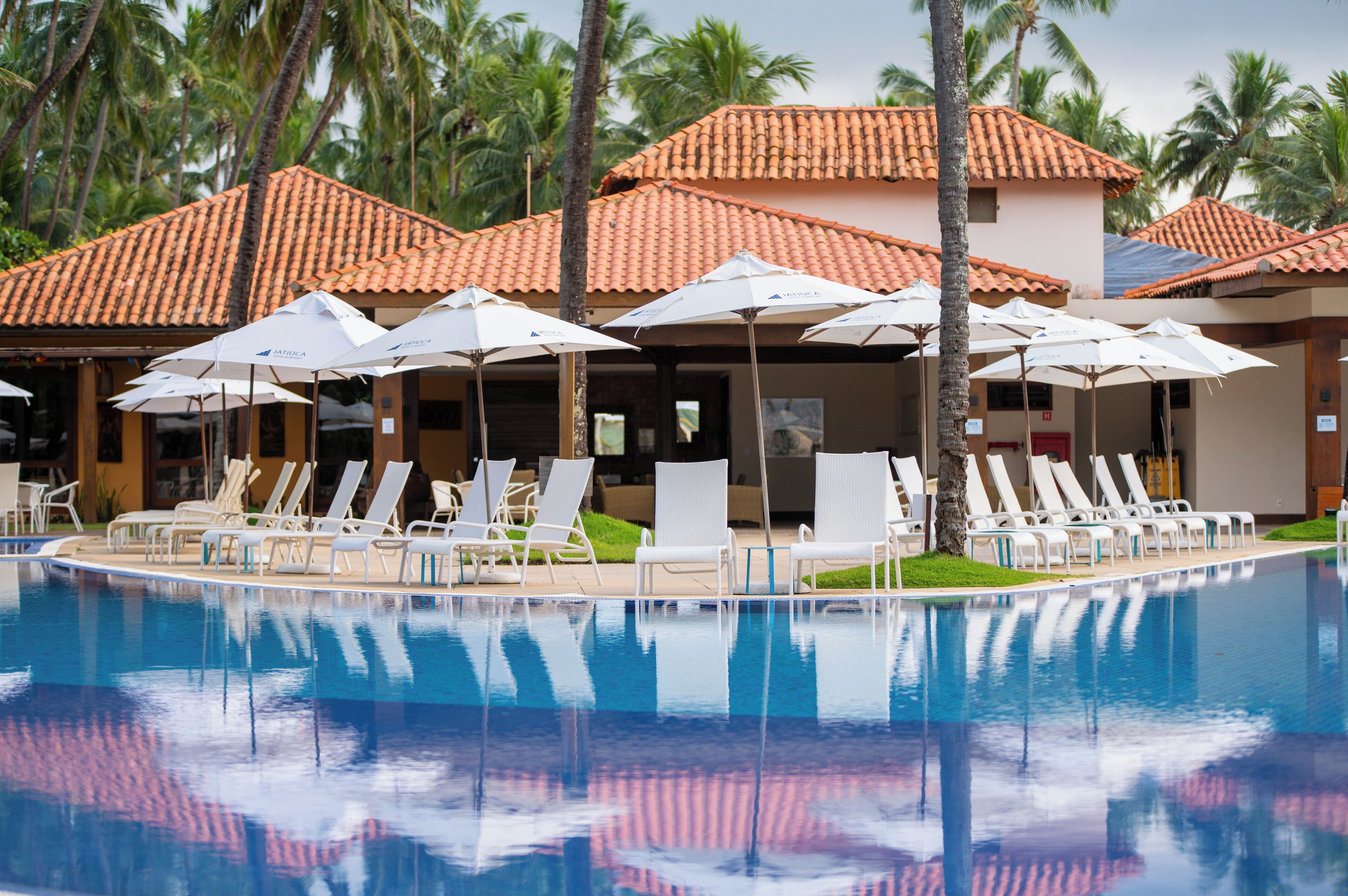 A pool area with lounge chairs and umbrellas, surrounded by palm trees and buildings with tiled roofs.
