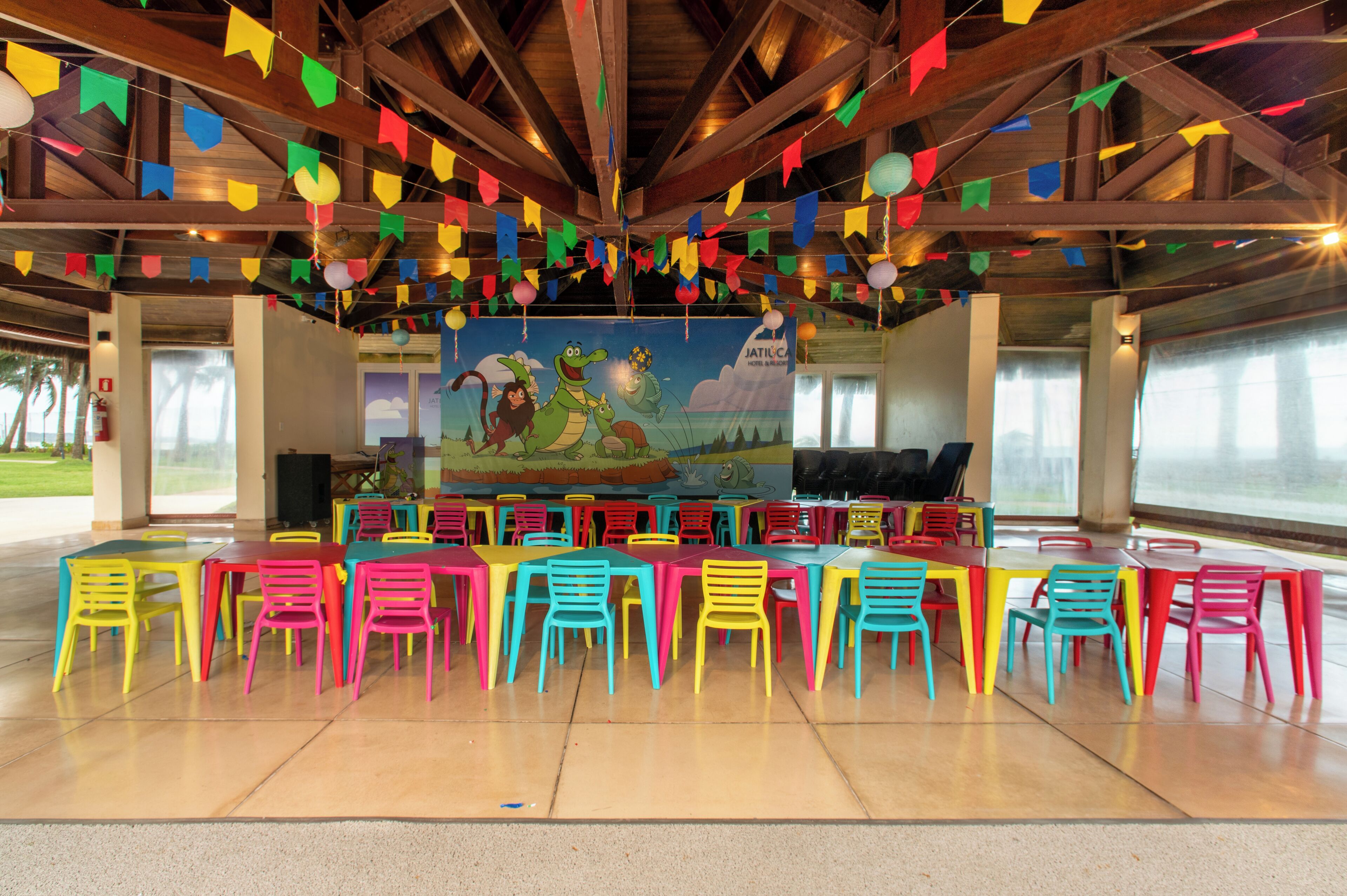A covered outdoor dining area with colorful plastic chairs and tables, a mural on the back wall, and a view of the outdoors.