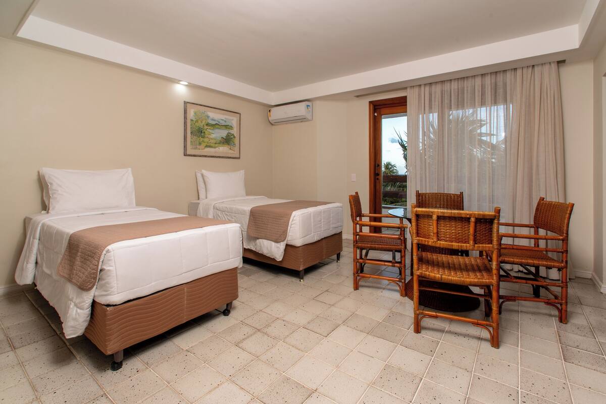 A hotel room with two beds, a dining table with chairs, a painting on the wall, and an air conditioning unit.