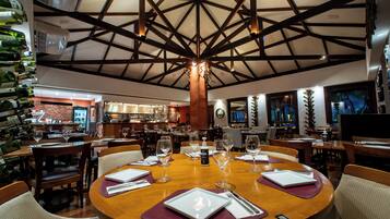 Un restaurante con techo alto y vigas a la vista, mesas de madera con copas de vino y cubiertos, y una zona de bar con botellas.