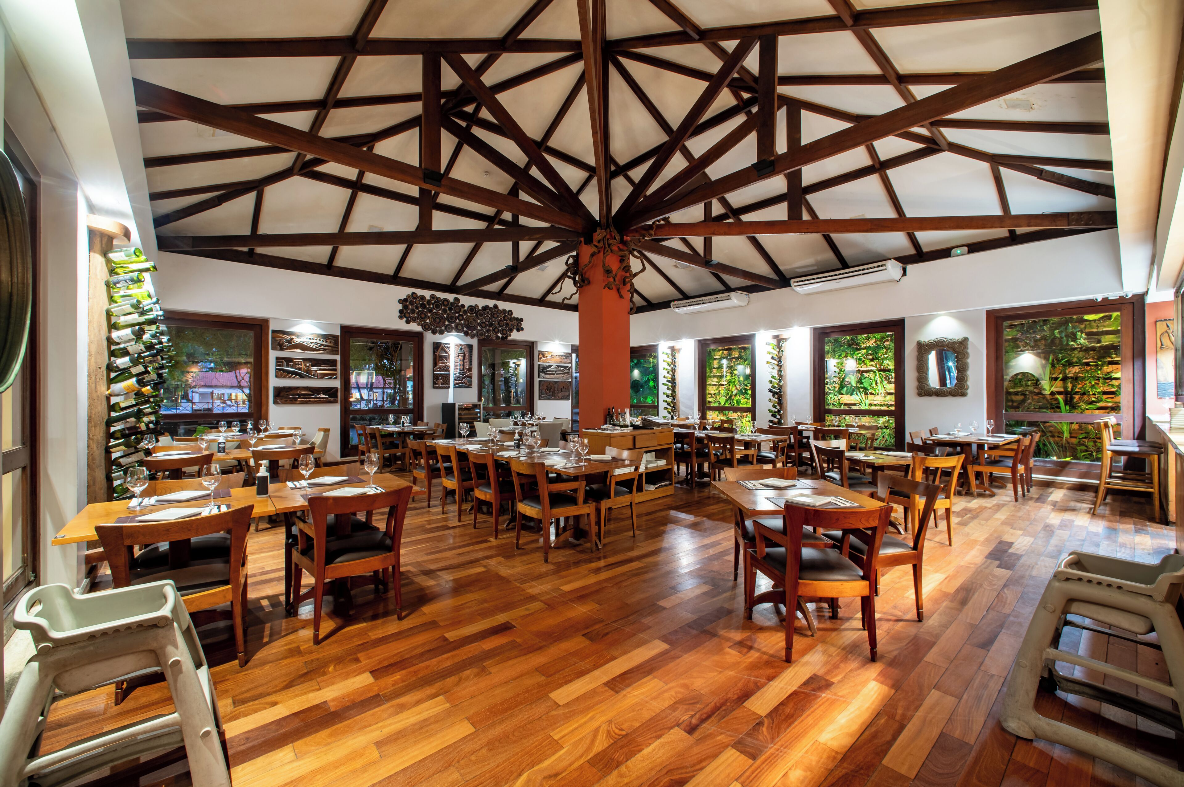 Un restaurante espacioso con mesas y sillas de madera, techo alto con vigas a la vista y ventanas amplias desde las cuales se ve follaje exterior.