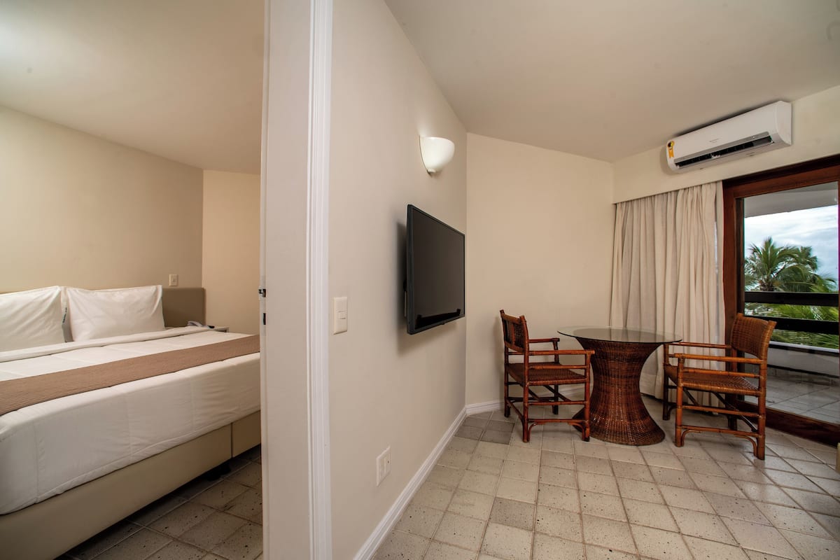 A hotel room with a bed, a TV, a dining table with chairs, and a balcony with a view.