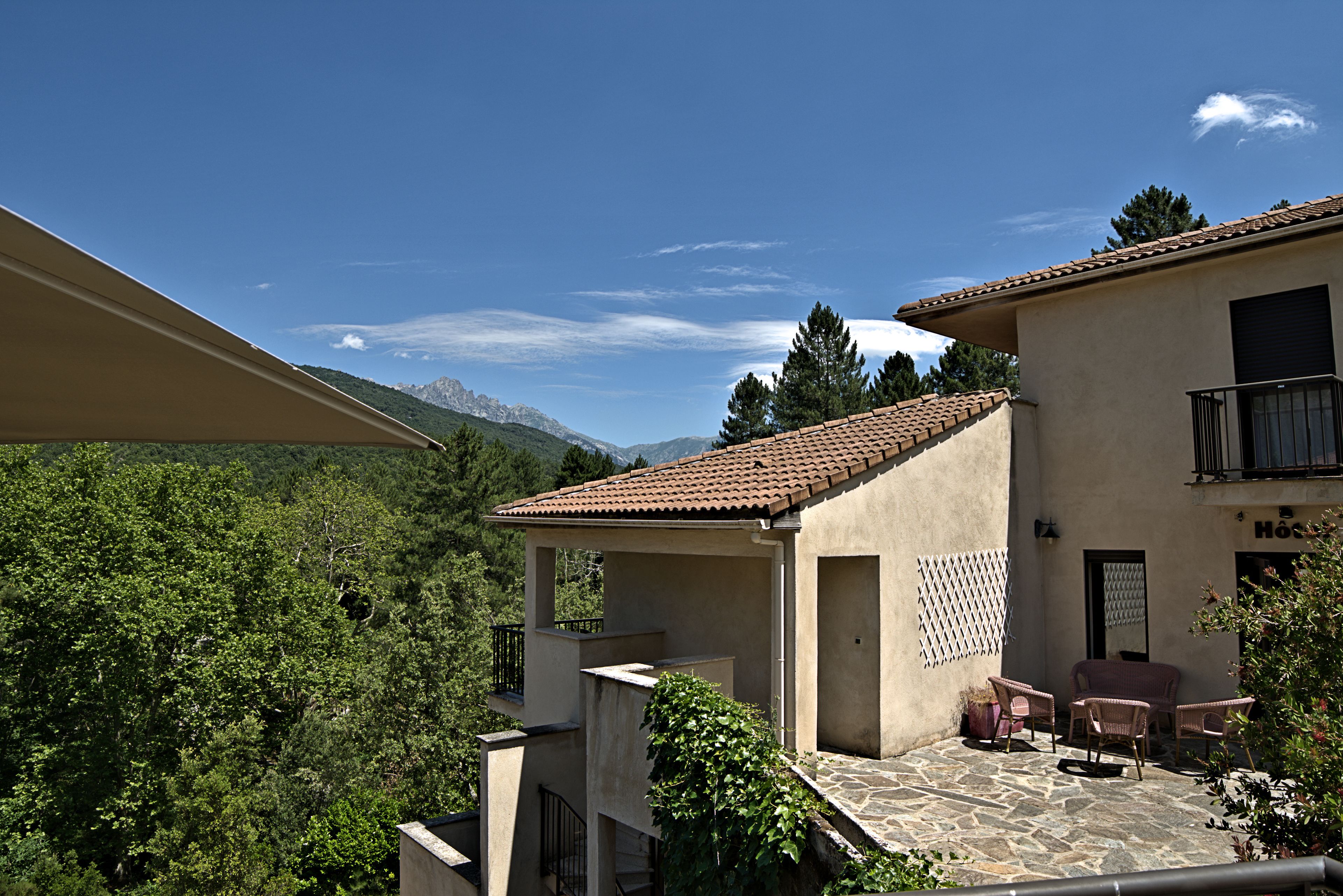 Un bâtiment à deux étages, doté d’un toit en tuiles, d’un balcon avec des meubles en osier et offrant une vue sur une chaîne de montagnes boisée.