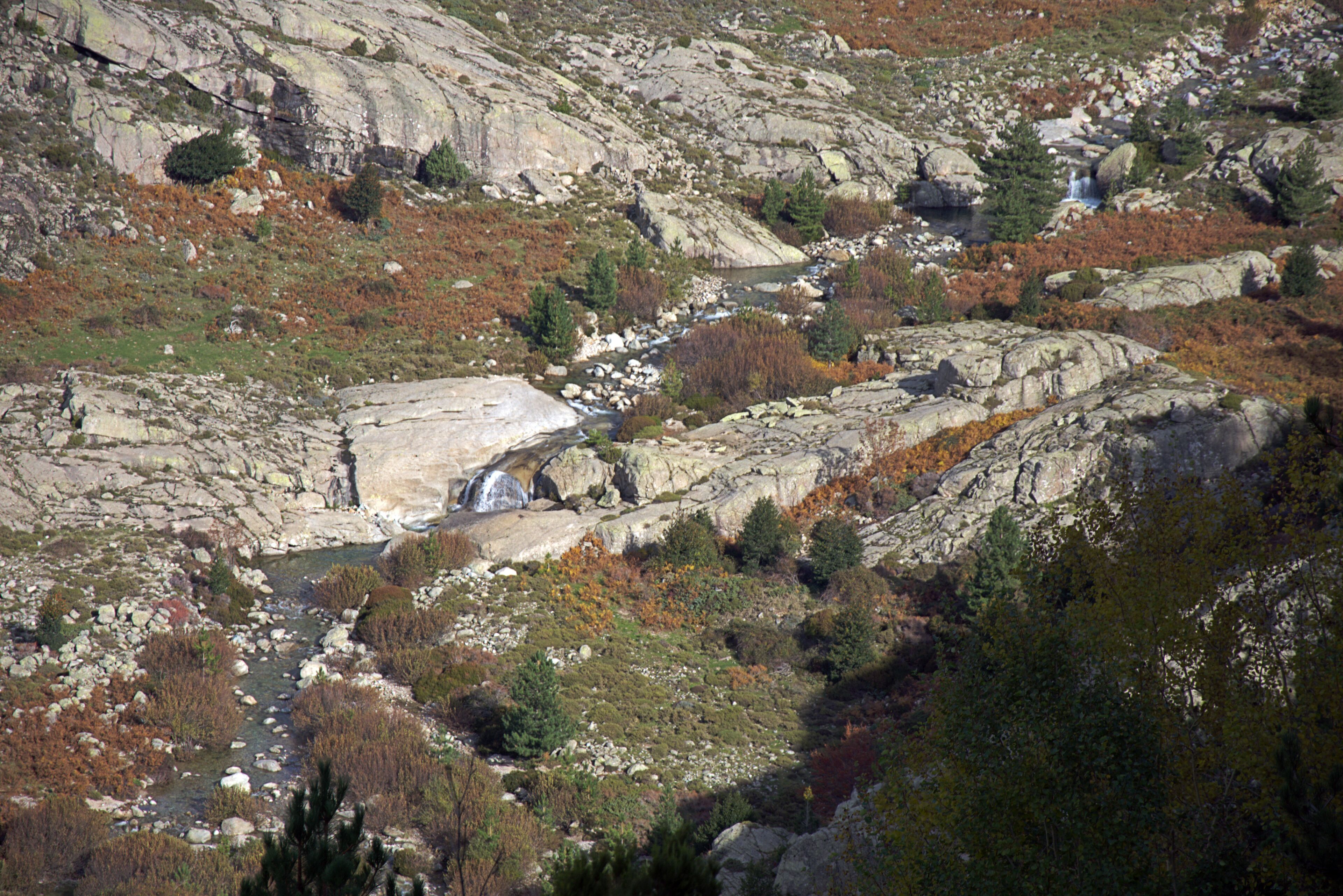 A mountainous landscape with a rocky stream, waterfalls, and sparse vegetation.