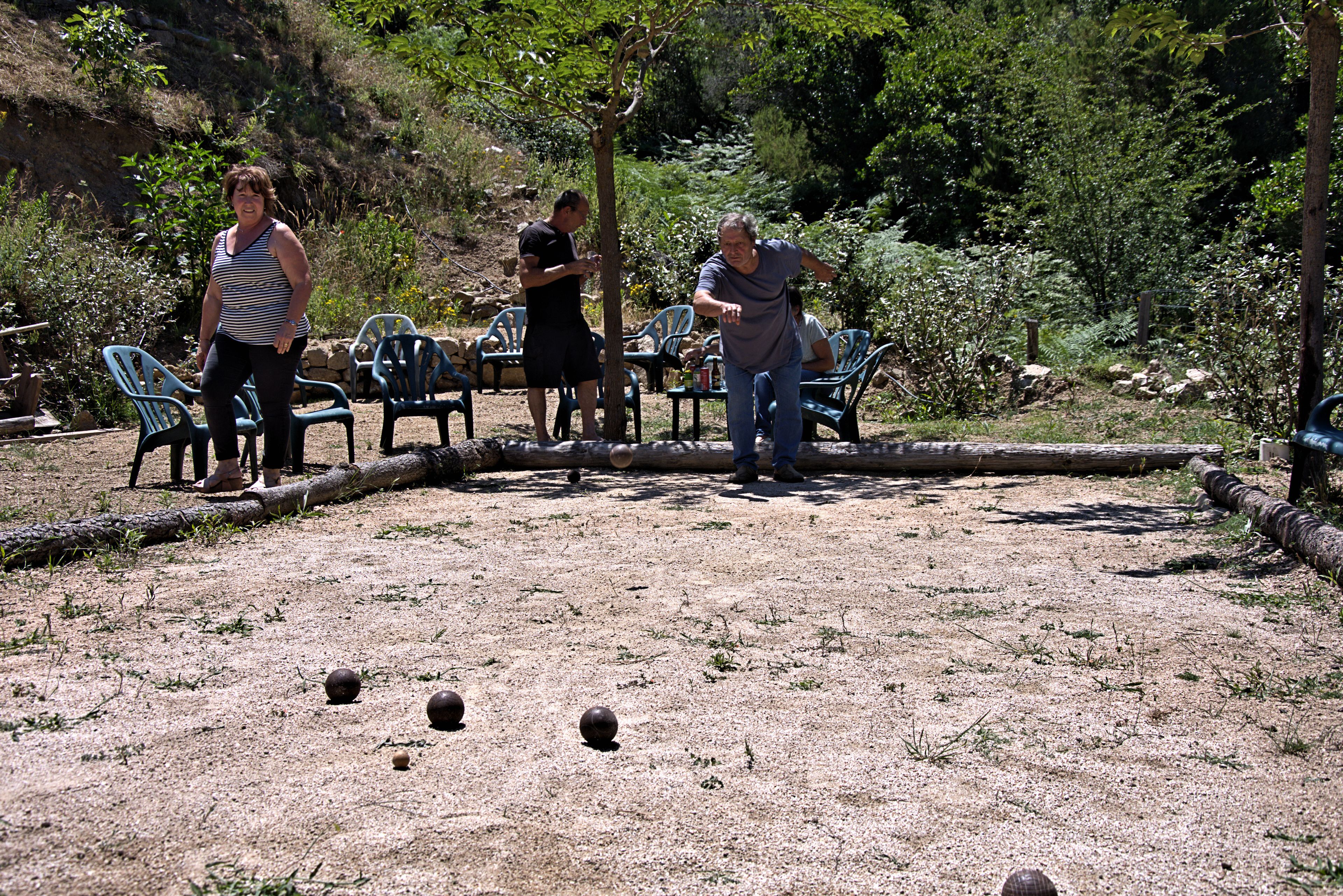 A group of people playing a game with balls on a gravel surface, surrounded by trees and greenery.