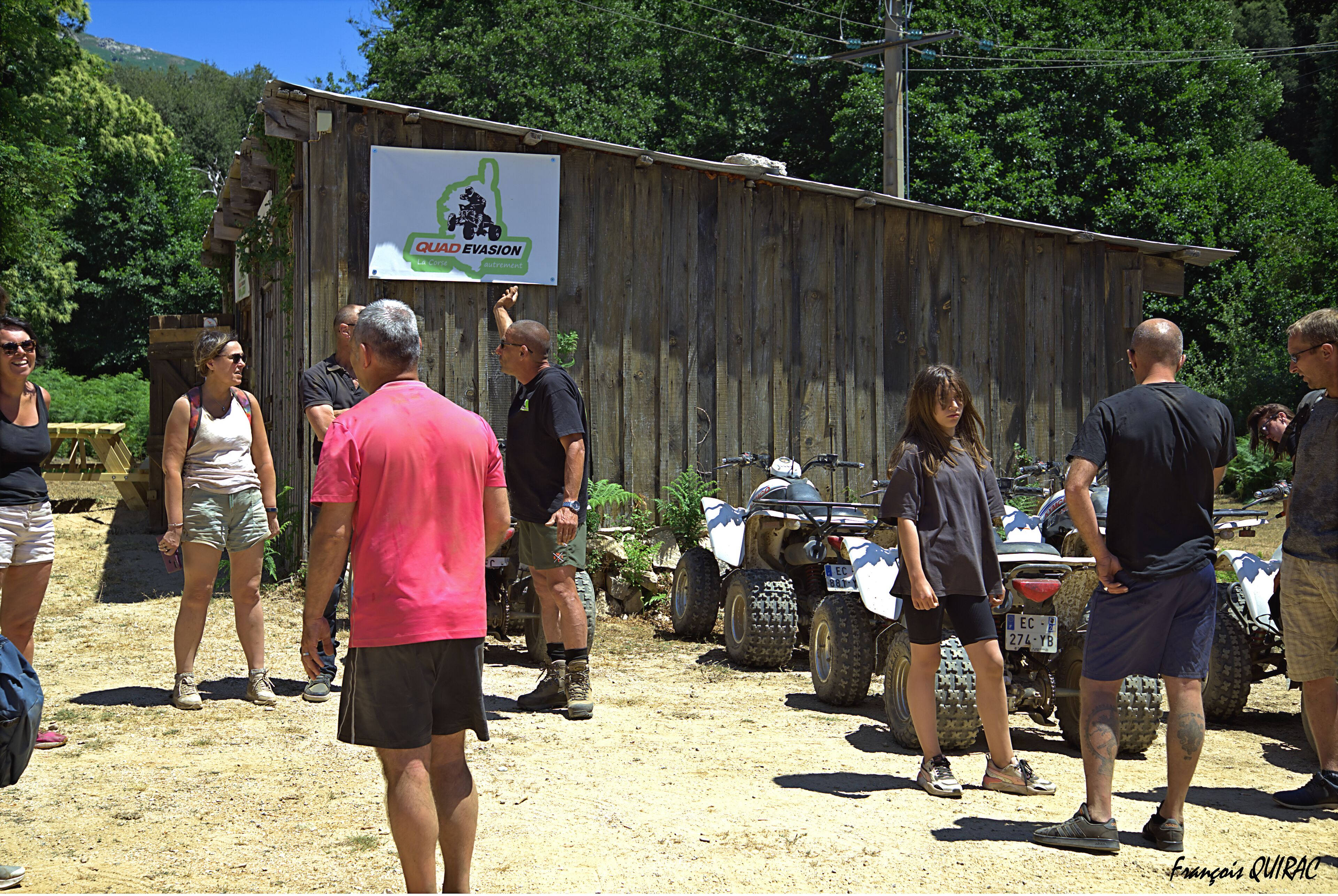 Un groupe de personnes se tient à l’extérieur, près d’un bâtiment en bois sur lequel on peut lire l’inscription : « QUAD EVASION ».