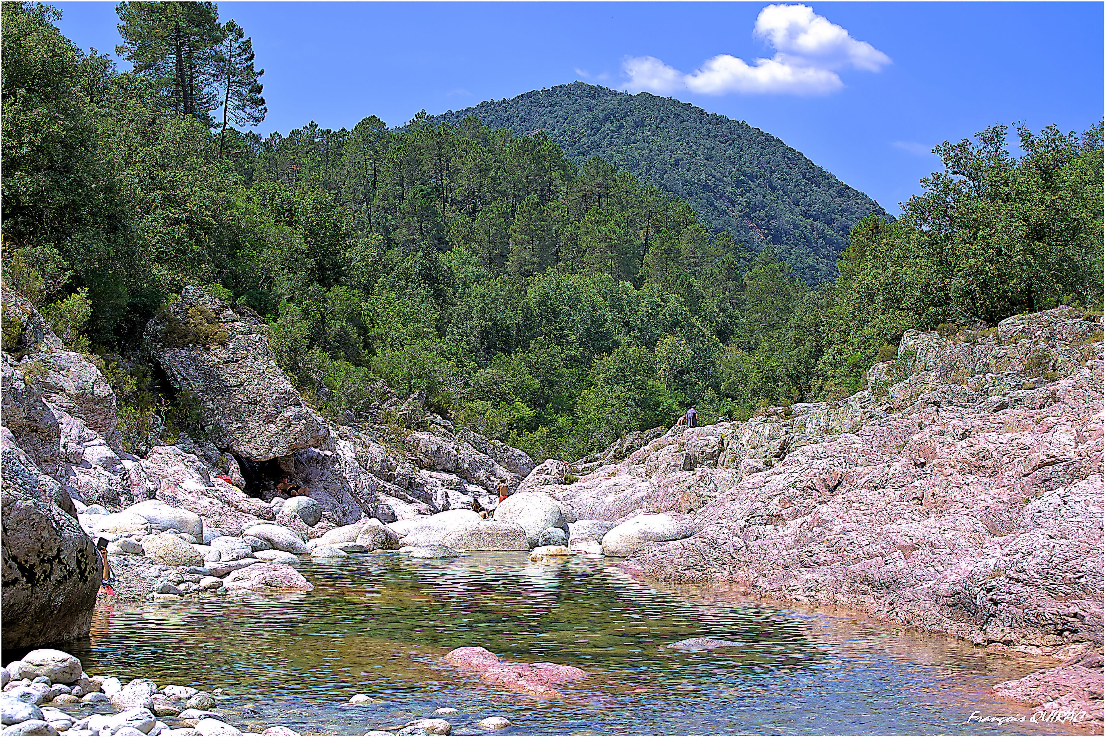 Un paisaje natural con un arroyo claro, terreno rocoso y una montaña boscosa al fondo.