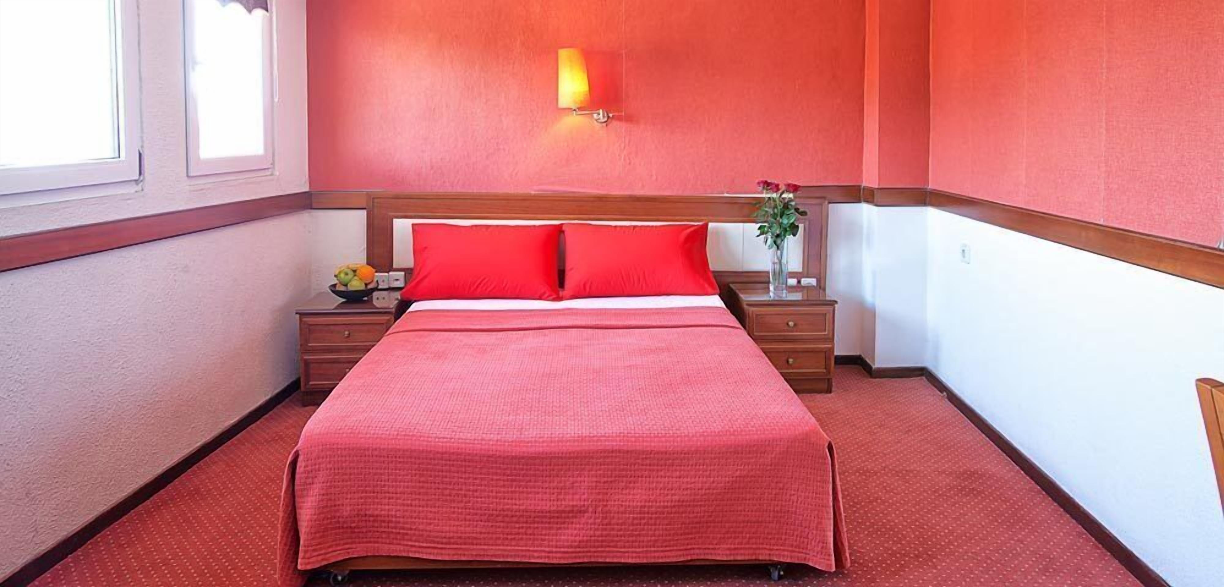 A neatly made bed with red bedding, a bedside table with a fruit bowl, and a wall-mounted lamp.