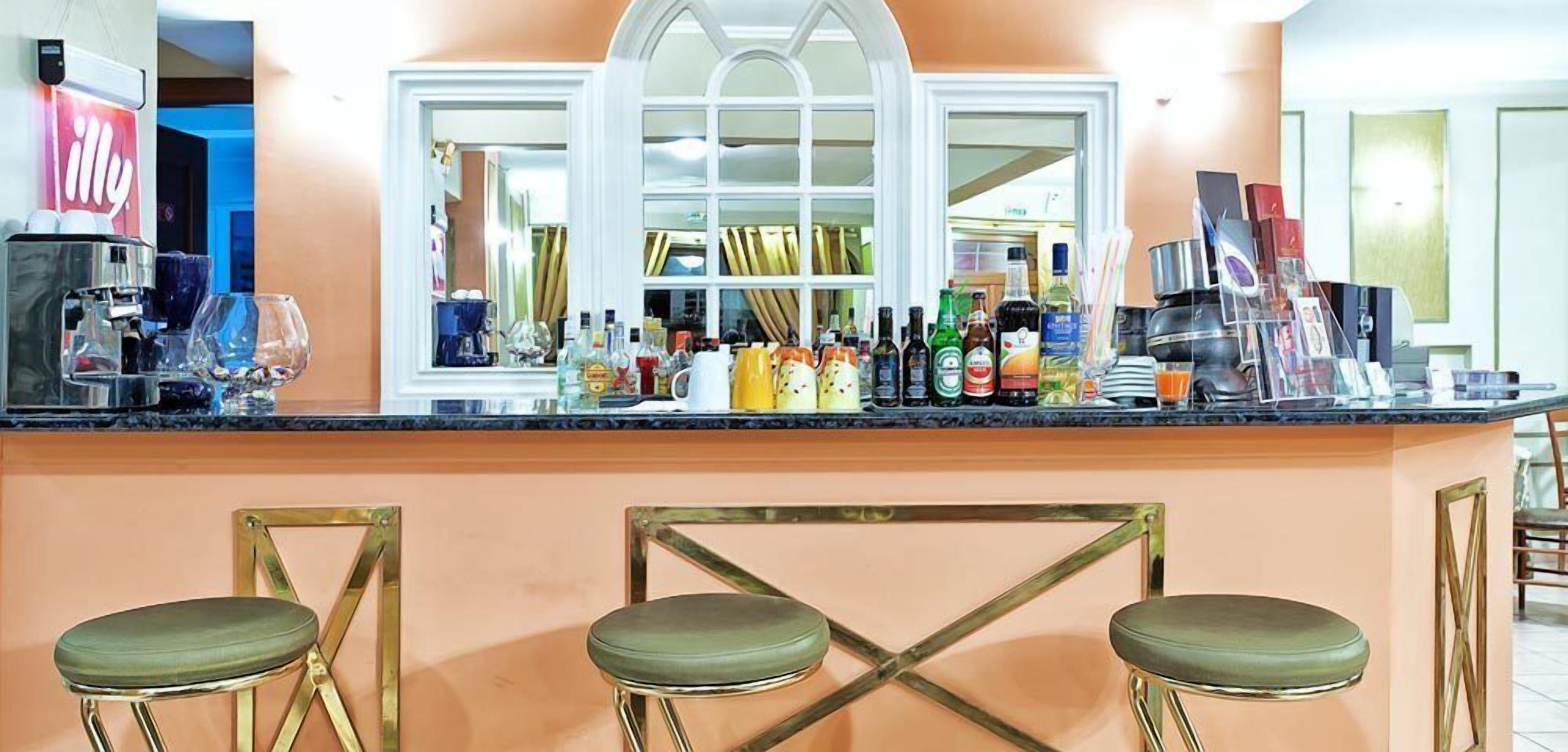 Zona de bar con una máquina de café, varias botellas y una ventana con cortinas. Hay tres taburetes con asientos verdes.