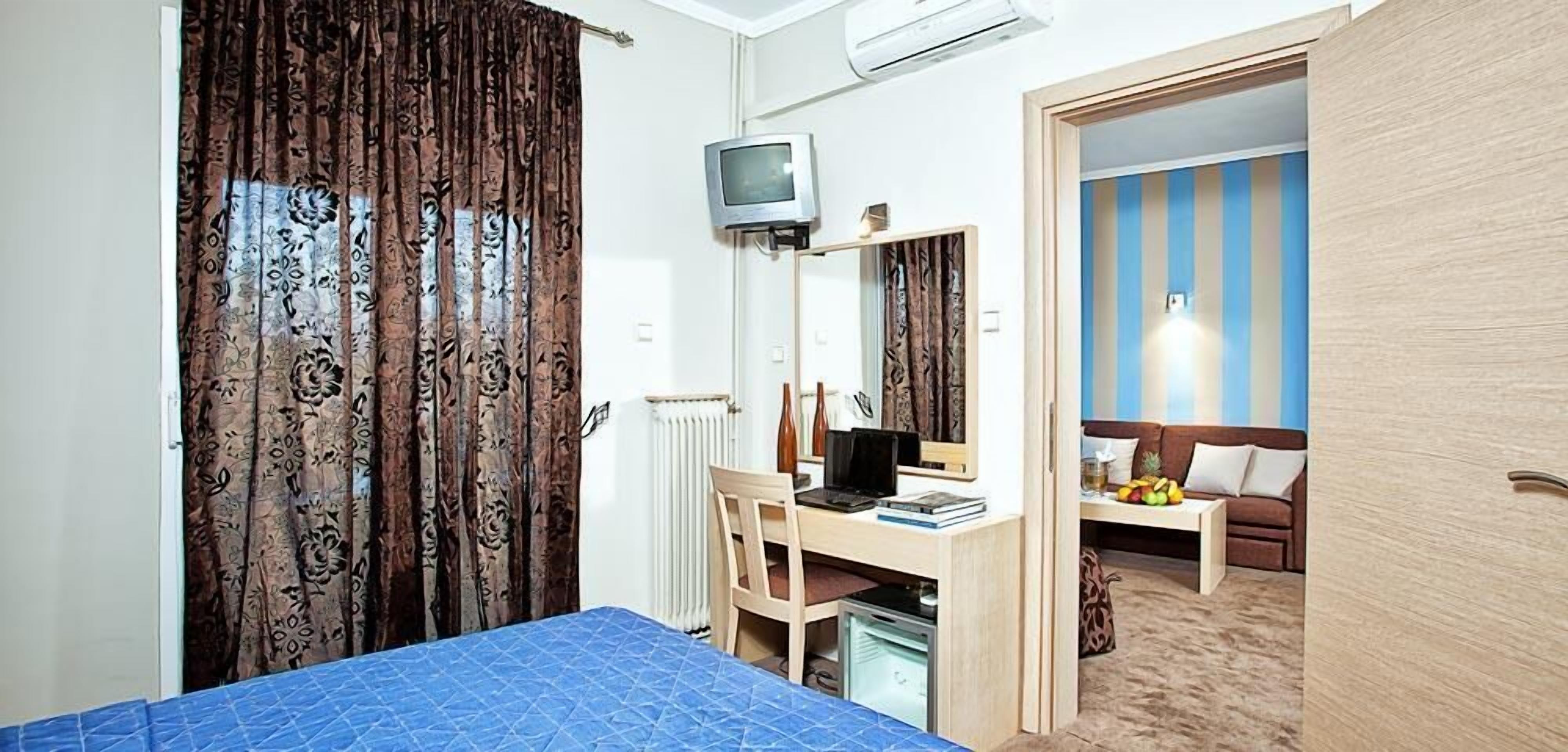 A bedroom with a bed, a desk with a computer, a TV, and a mirror. There is a window with curtains and a radiator.
