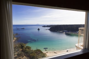 Vista costera desde una ventana, con mar azul turquesa, playa arenosa y acantilados rocosos.