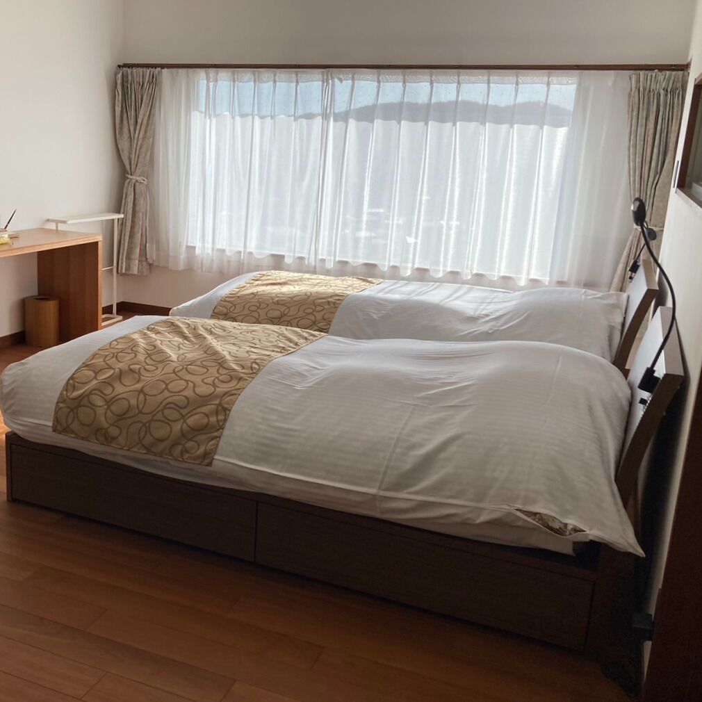 Una cama doble con colcha estampada, una mesita de noche, una lámpara y una ventana con cortinas translúcidas.