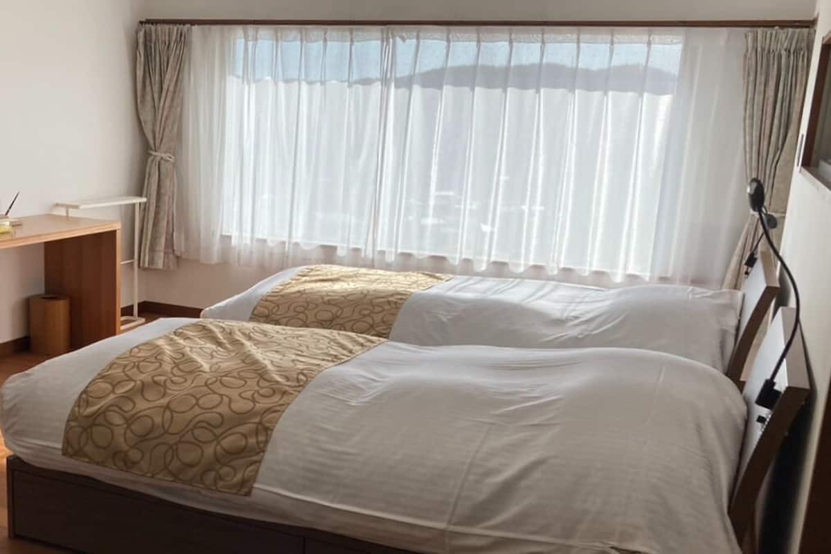 Una cama doble con colcha estampada, una mesita de noche, una lámpara y una ventana con cortinas translúcidas.