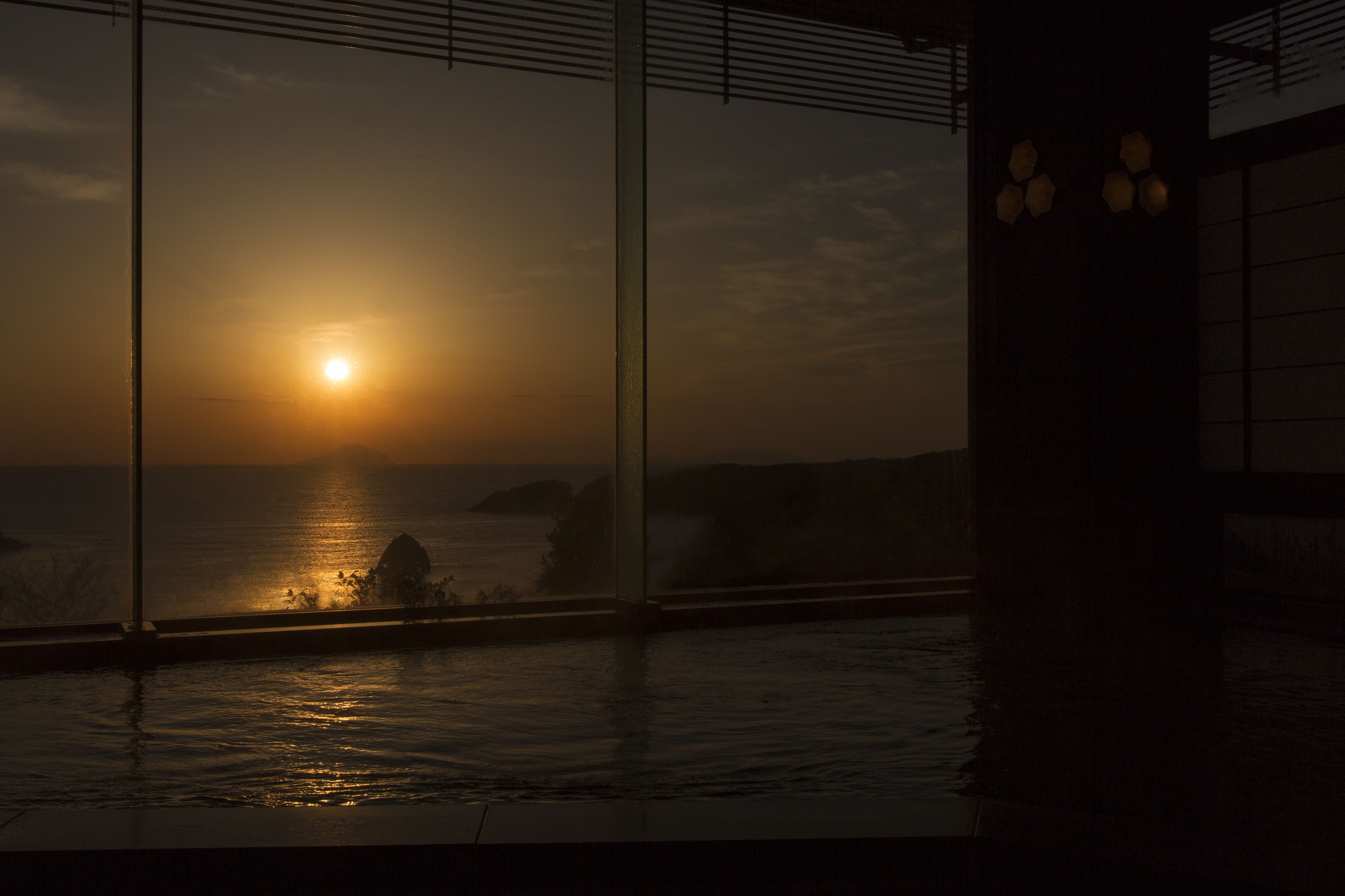 A sunset view through a window overlooking a pool, with a clear sky and distant landforms.