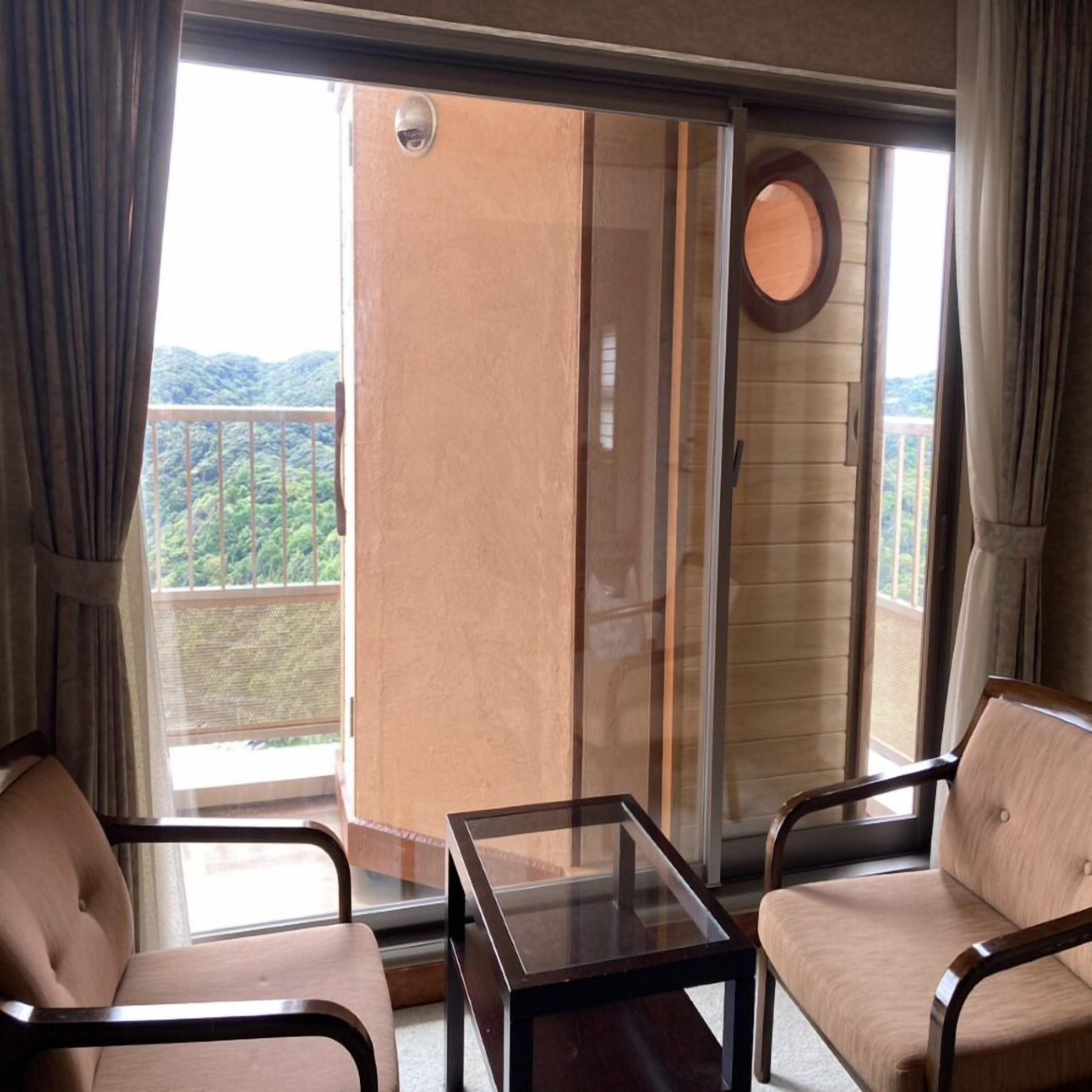 A balcony with a glass door, two chairs, a small table, and a view of a mountainous landscape.