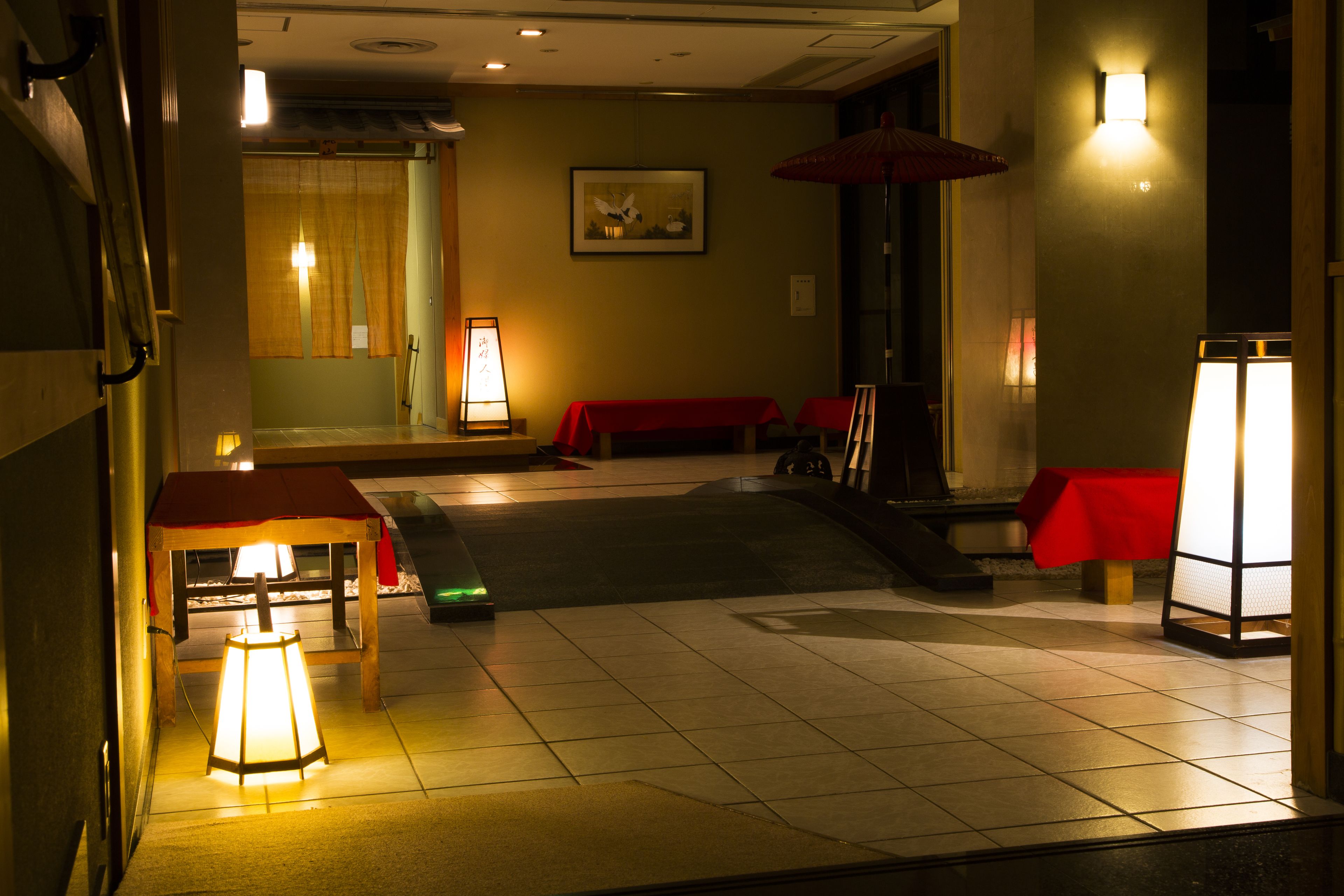 Un vestíbulo de hotel con piso de baldosas, mesas rojas y algunas sillas. Hay luces fijadas a la pared y un cuadro enmarcado.