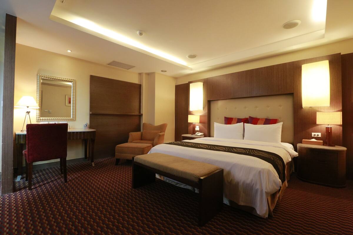 A hotel room with a large bed, a desk, a chair, a bench, and two bedside lamps.