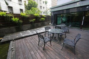 Un patio al aire libre con suelo de madera, sillas y mesas de metal. Se observa una pared con plantas verdes y una pequeña fuente de agua.