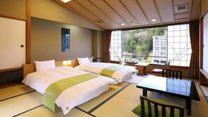 A hotel room with two beds, a small table, and a view of a building through large windows. - Katsuragawa (Izu)