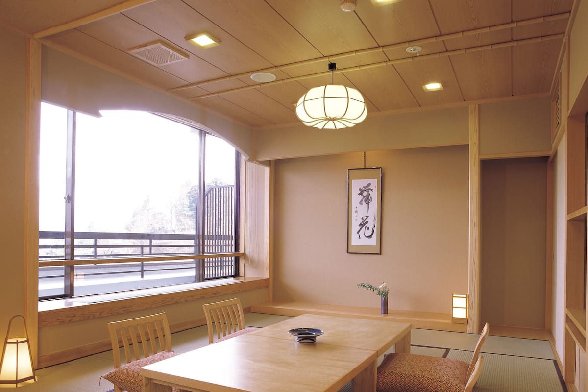 A traditional Japanese room with tatami flooring, a low table, wooden chairs, a hanging light fixture, a window with a view, and a framed calligraphy piece on the wall.