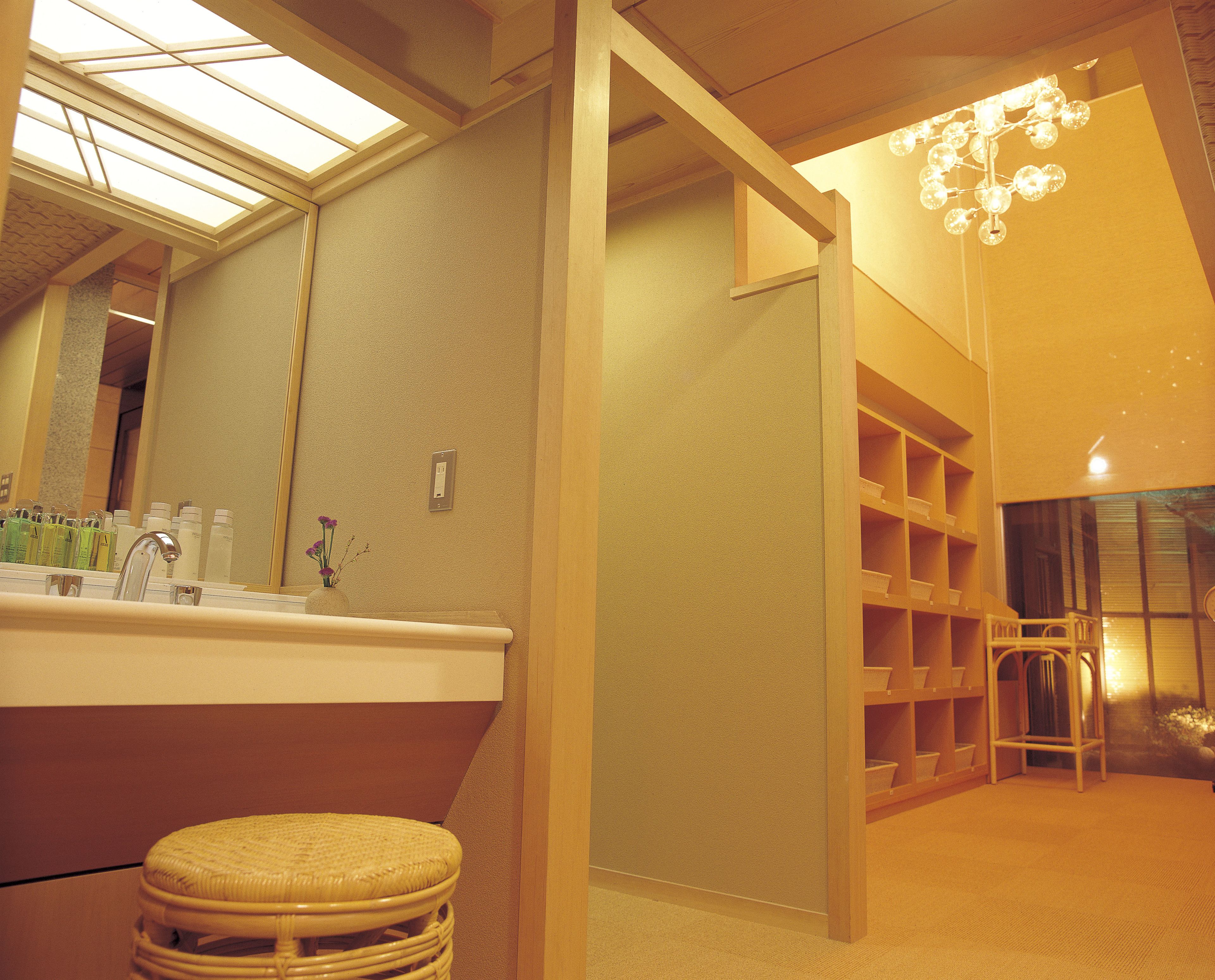 A bathroom with a large mirror, a sink, and a wicker stool. There is a shelf with toiletries and a chandelier.