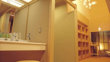 A bathroom with a large mirror, a sink, and a wicker stool. There is a shelf with toiletries and a chandelier.