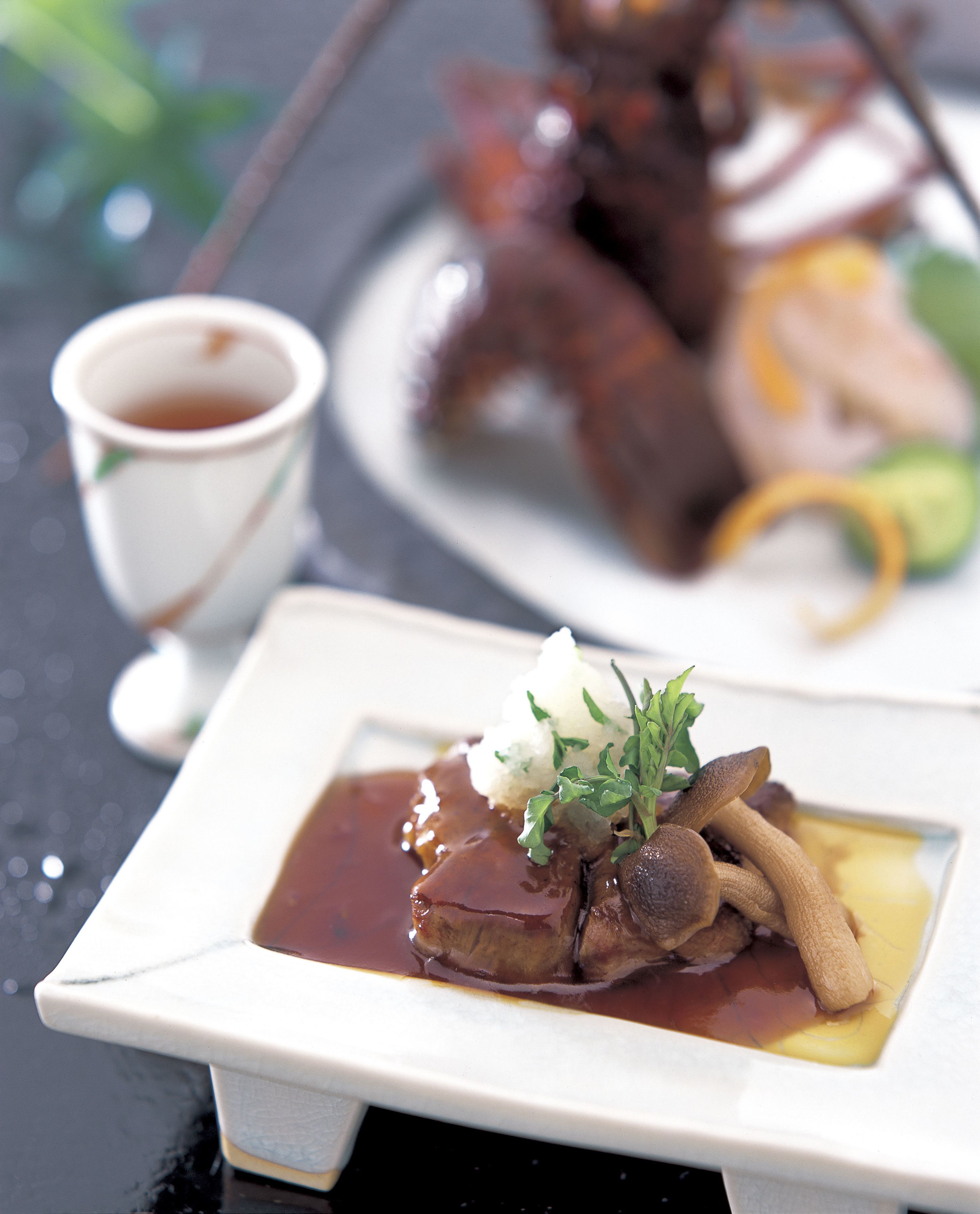 Un plato cuadrado con guiso de carne, salsa y decoración, acompañado de una taza pequeña de salsa y un plato con carne en rodajas al fondo.