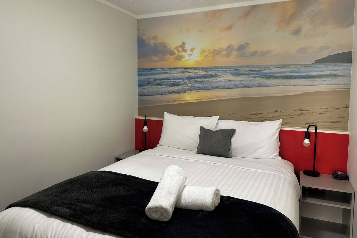 Ein Hotelzimmer mit Bett, roter Wand, Nachttisch mit Lampe und einer großen Strandmalerei an der Wand.